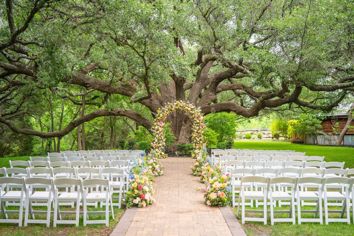 Pastel floral arch and aisle arrangements beneath a sprawling oak, white chairs set on the lawn