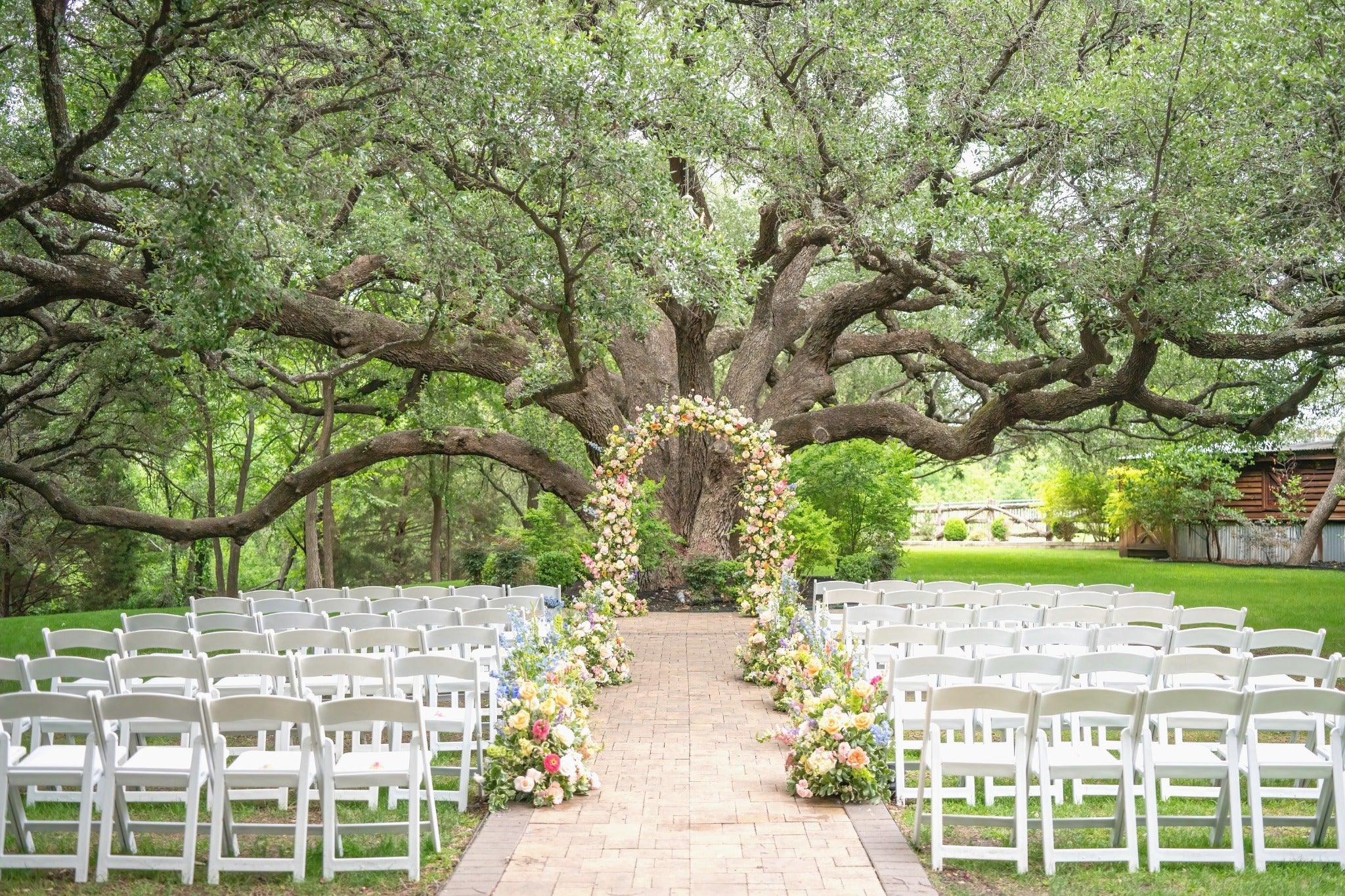 Outdoor wedding setup with white chairs and floral arch under a large tree.
