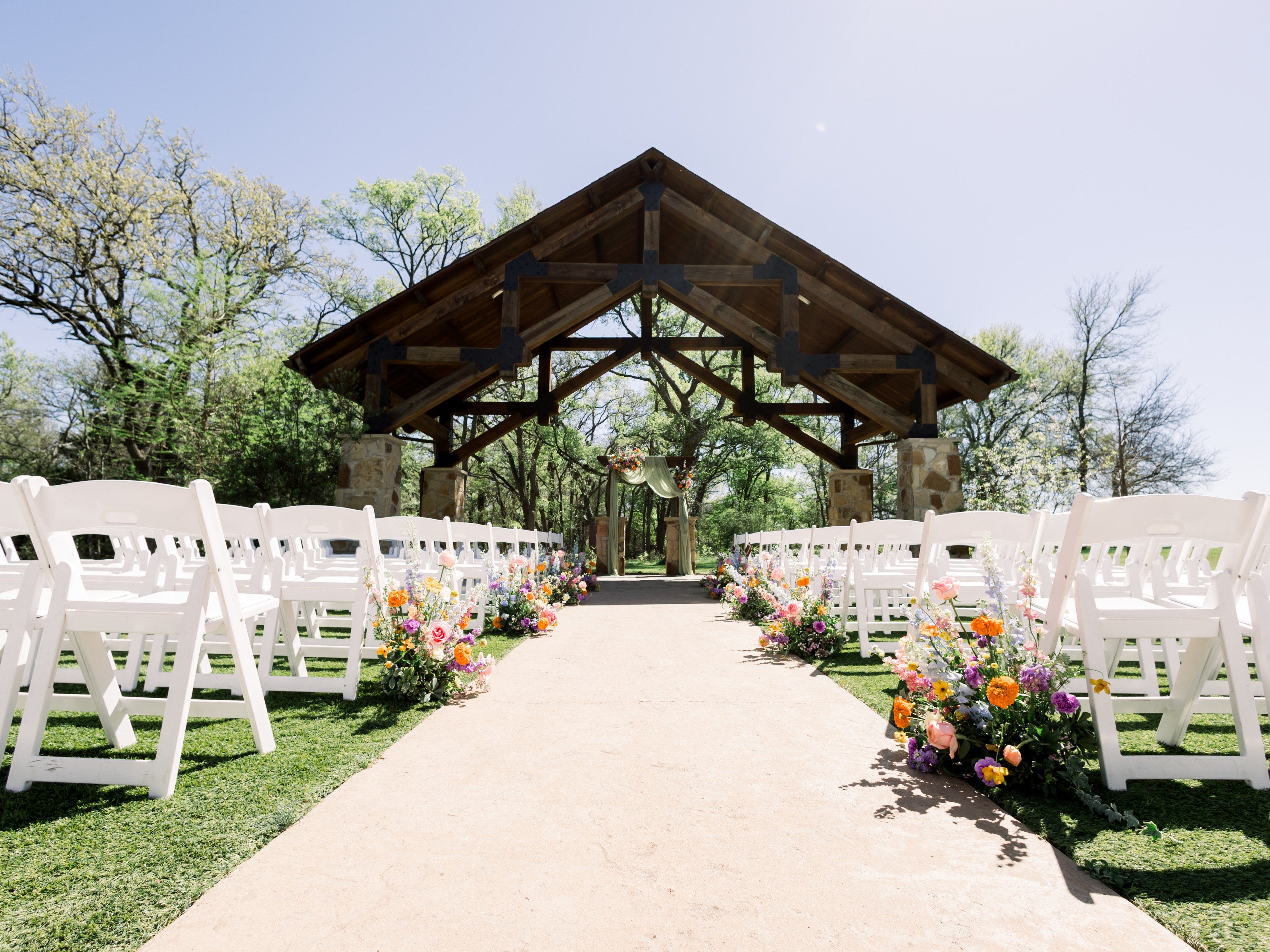 The wood-beam pergola at The Springs Alvarado in early spring, bare oaks behind, the aisle lined with coral, orange and pink ground florals by B.blumen