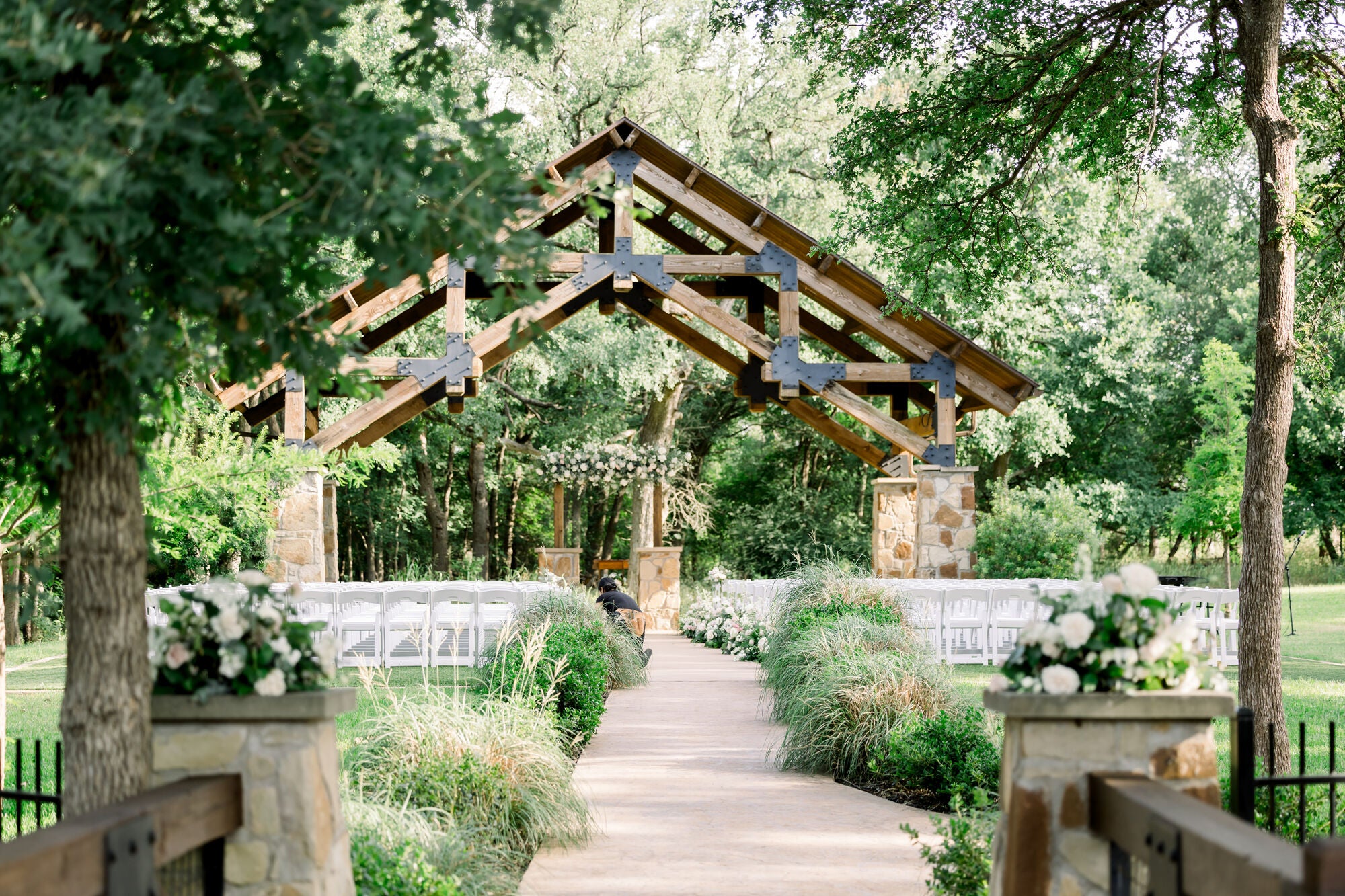 Approach to the pergola ceremony at The Springs Alvarado, stone gate piers in the foreground and the wood-beamed pergola altar in the distance, aisle lined with white florals by B.blumen