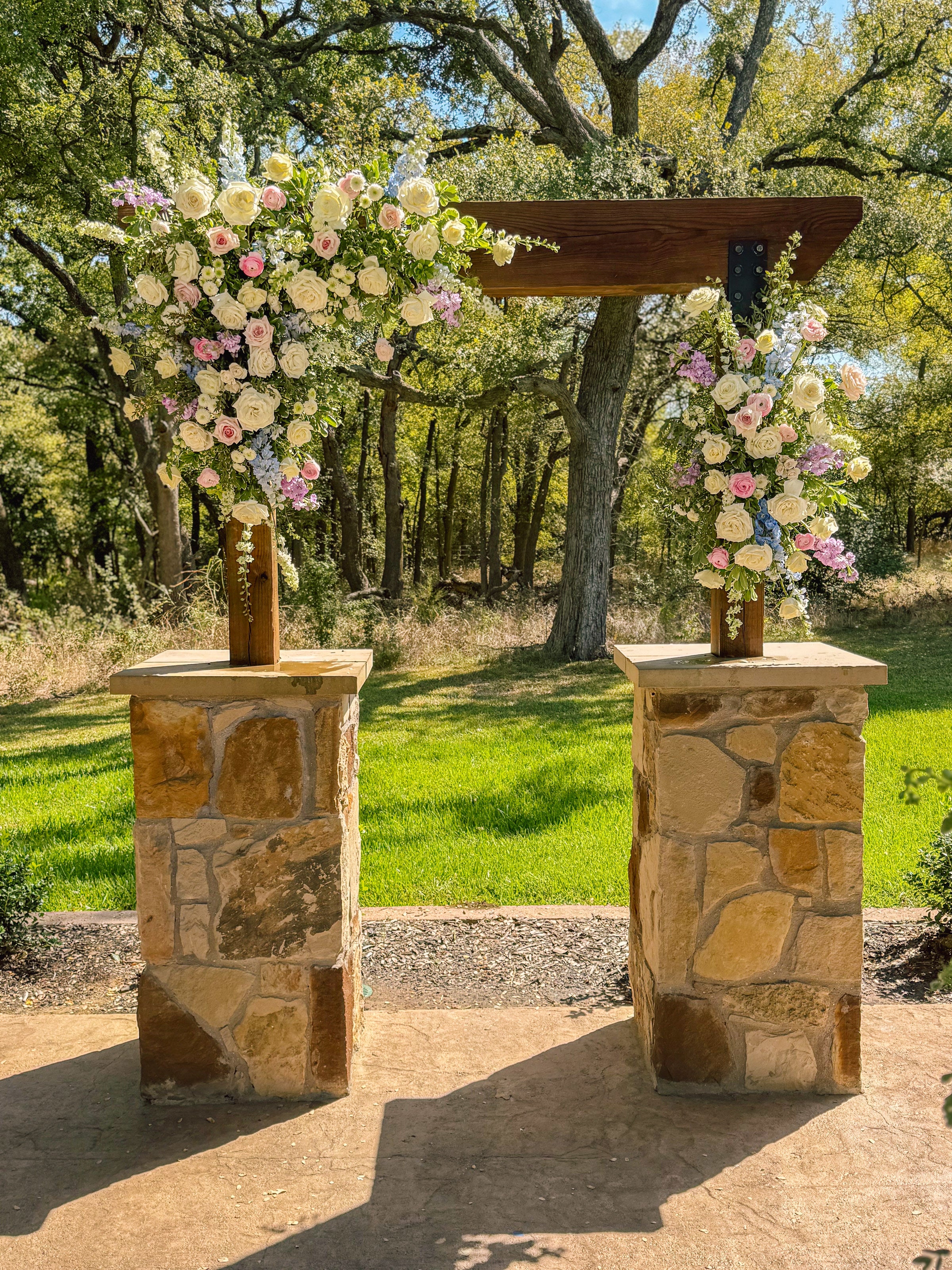 Two stone-pier pedestals at The Springs Alvarado topped with cascading pastel arrangements of white roses, blush, lavender, and pale blue blooms, beneath a wood-beam connector with the oak canopy behind, by B.blumen