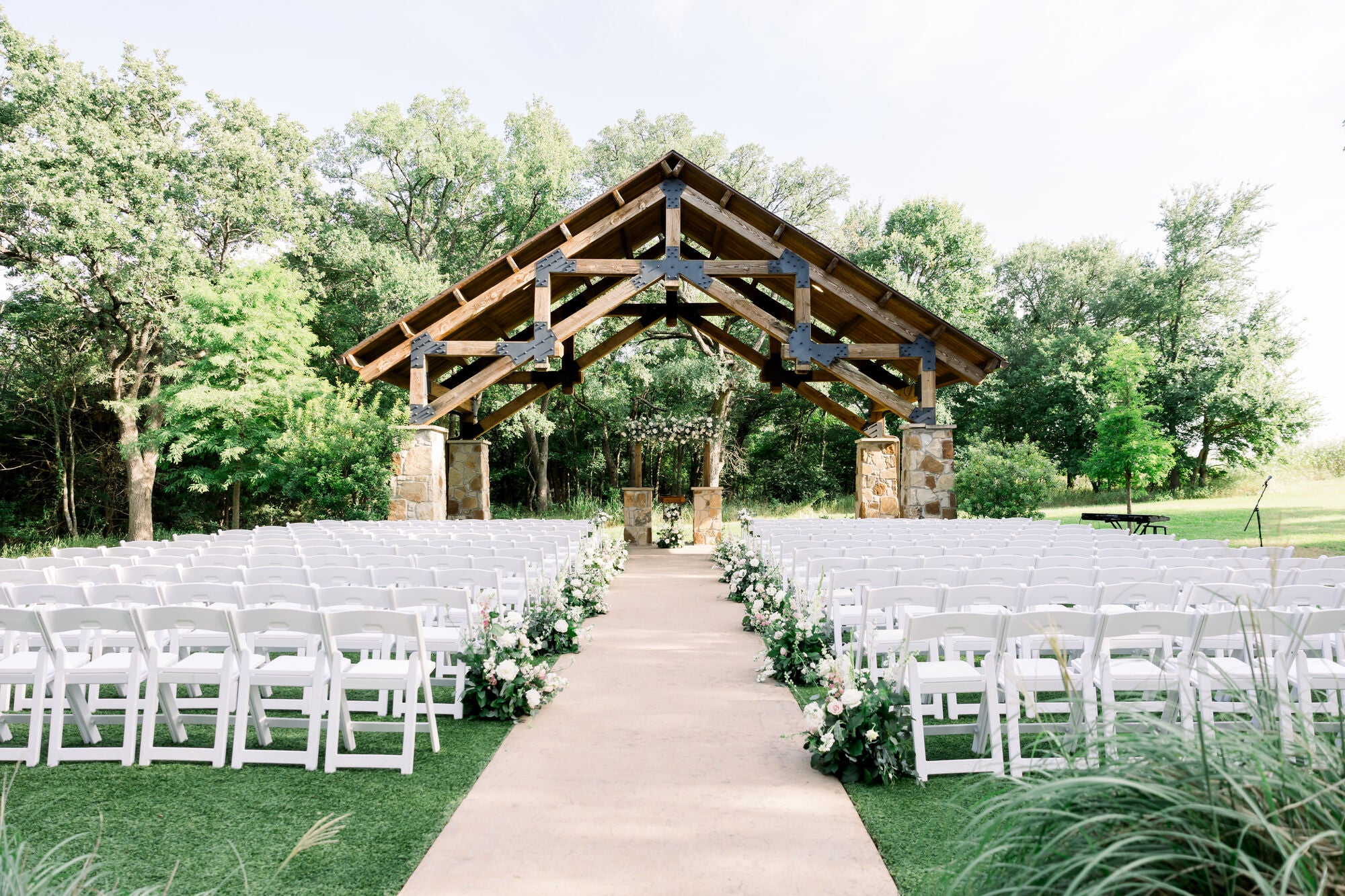 Wide view of the stone-and-wood pergola ceremony at The Springs Alvarado, brick aisle flanked by white pedestal florals and rows of garden chairs, framed by mature trees, by B.blumen