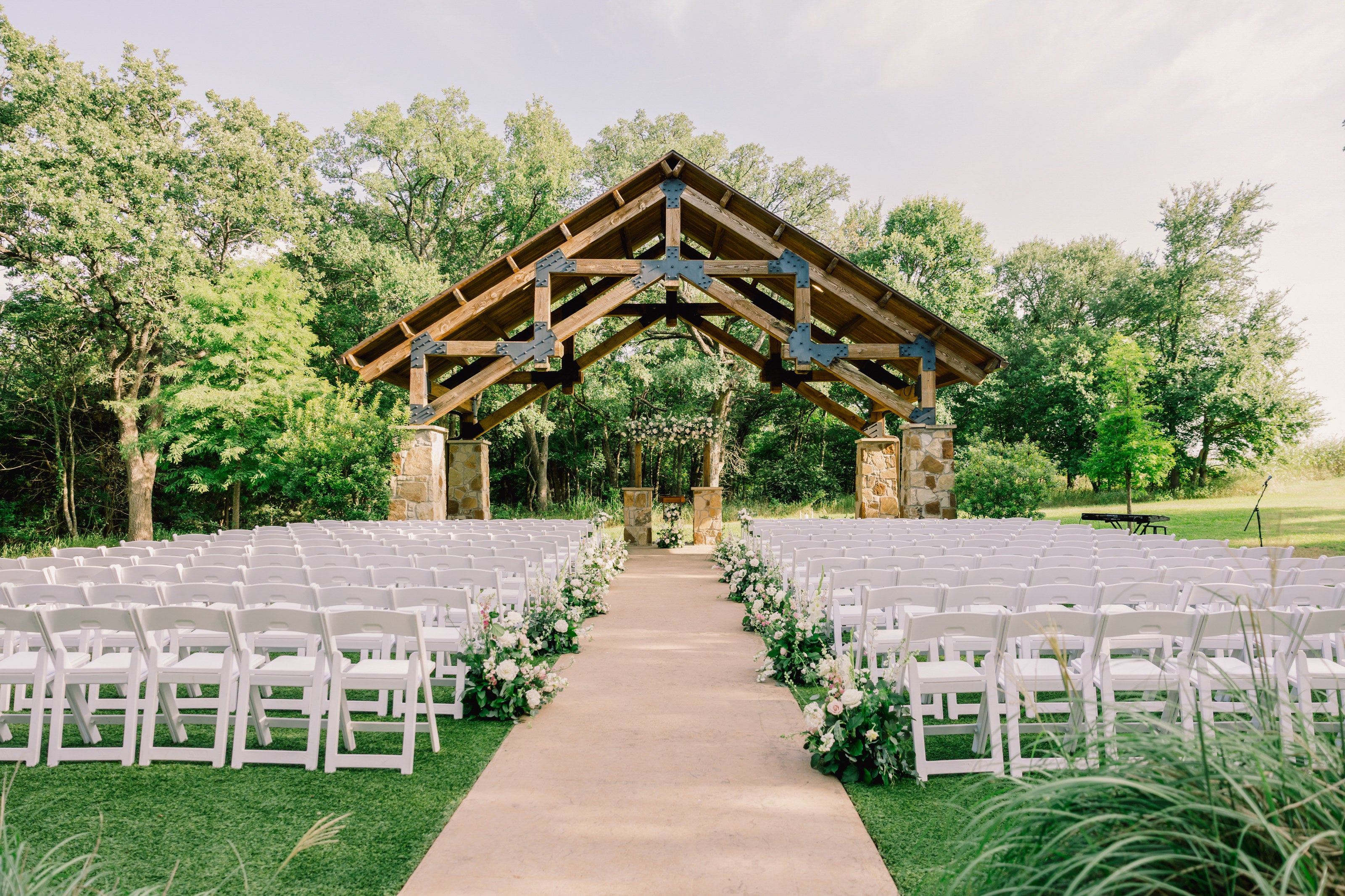 Wide view of the stone-and-wood pergola ceremony at The Springs Alvarado, a stone path lined with white and blush ground florals between rows of garden chairs, mature trees behind, by B.blumen