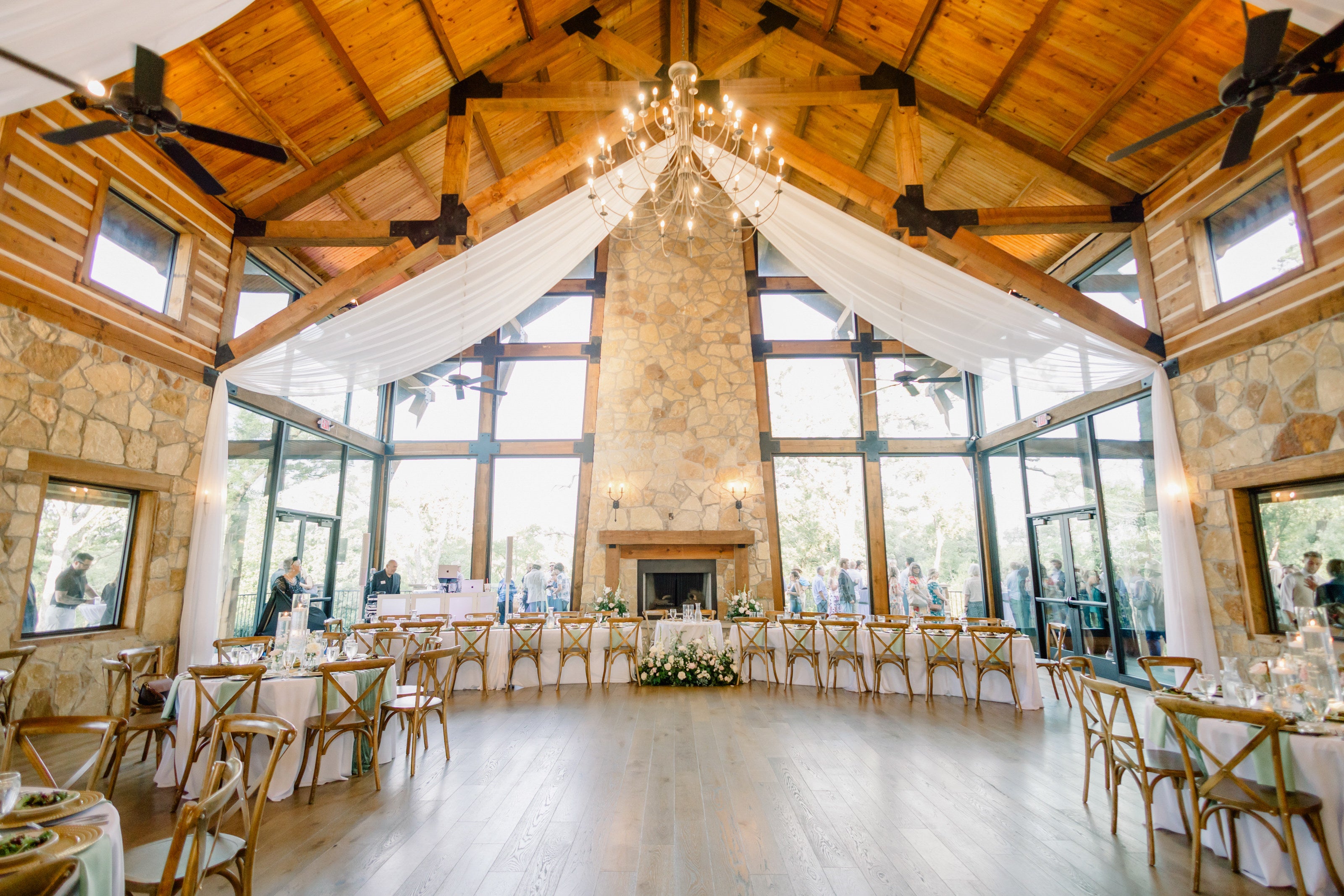 Interior of Timberview Lodge at The Springs Alvarado, cathedral ceiling with iron chandelier, three-story stone fireplace, draped fabric, and the head table set with low white centerpieces, by B.blumen