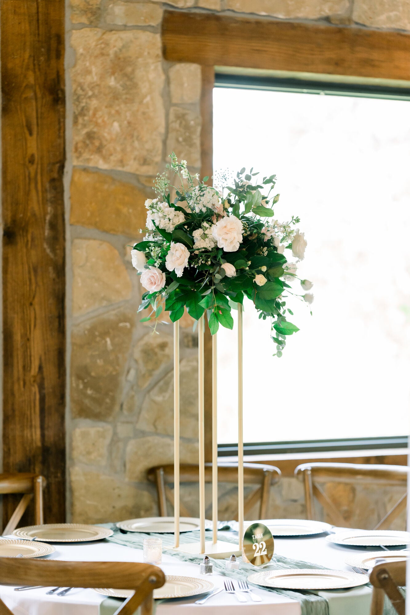 Tall pedestal centerpiece at Timberview Lodge with blush roses, white hydrangea, and trailing greenery, set against the stone wall and lodge window, by B.blumen