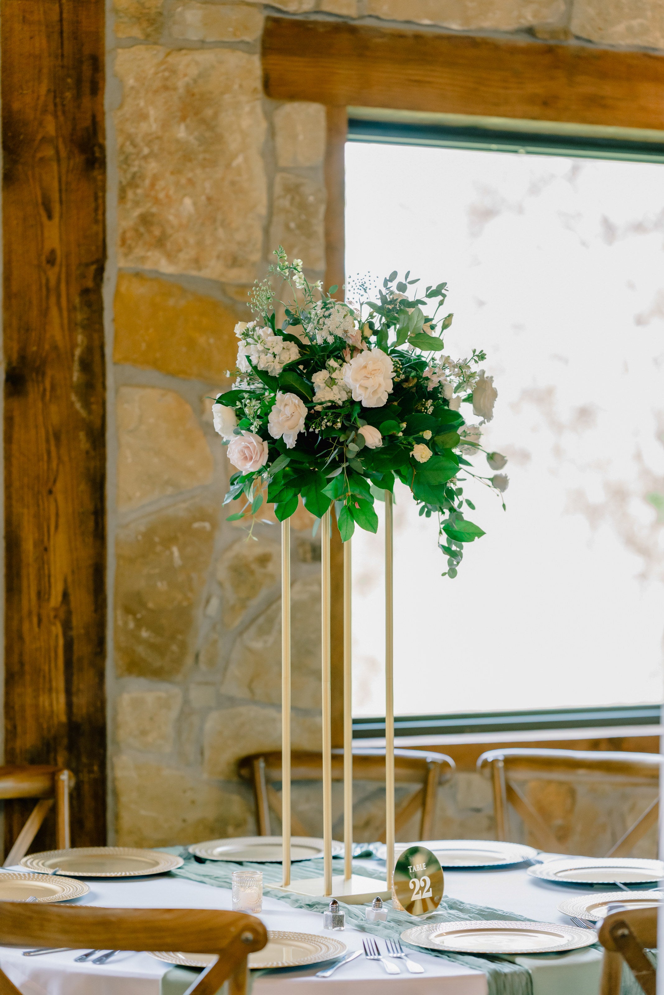 Tall pedestal centerpiece at Timberview Lodge with blush roses, white hydrangea, and trailing greenery, set against the stone wall and lodge window, by B.blumen