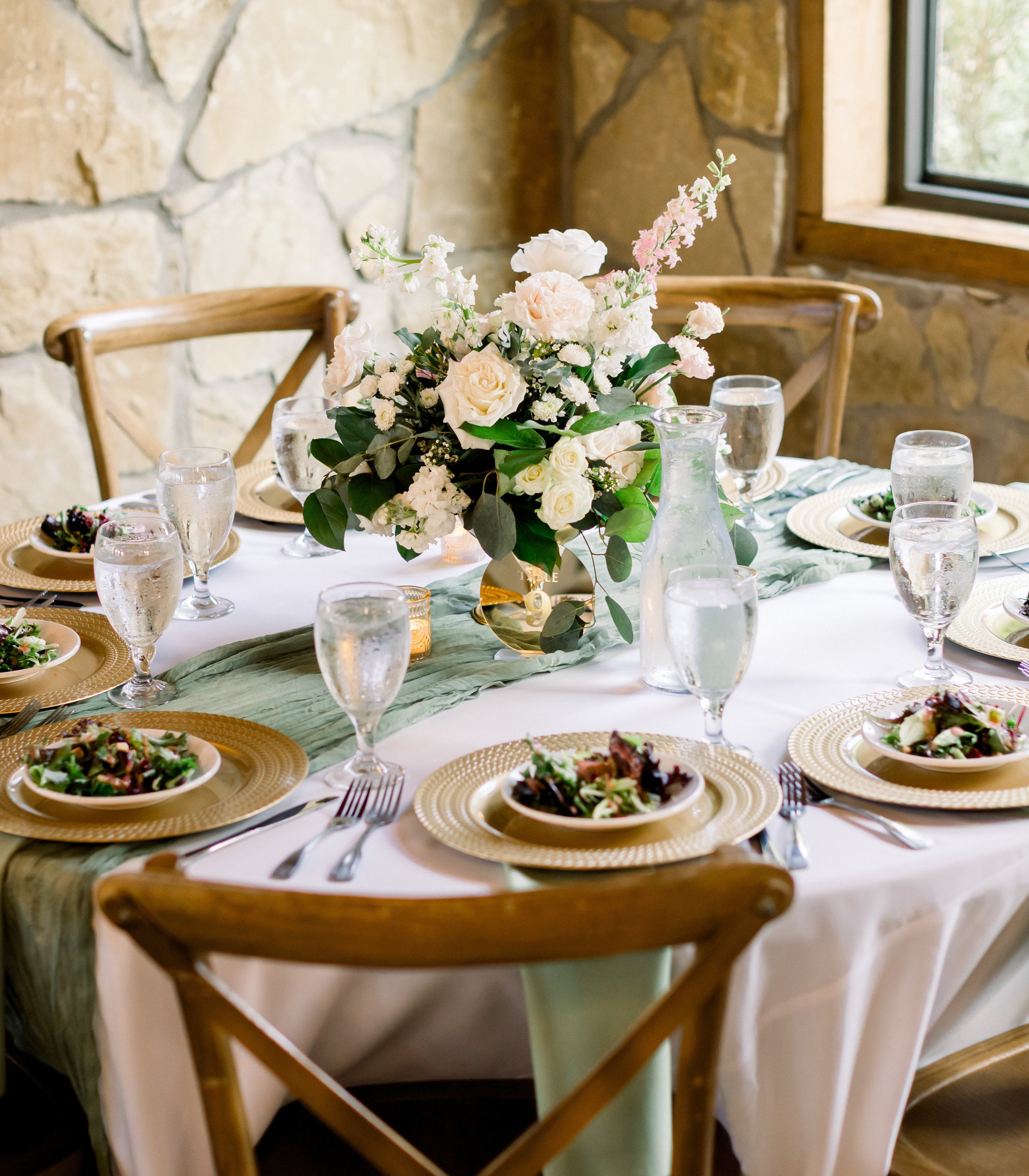 Round reception table at Timberview Lodge with a sage runner, gold chargers, and a low white-and-blush centerpiece against the stone wall, by B.blumen