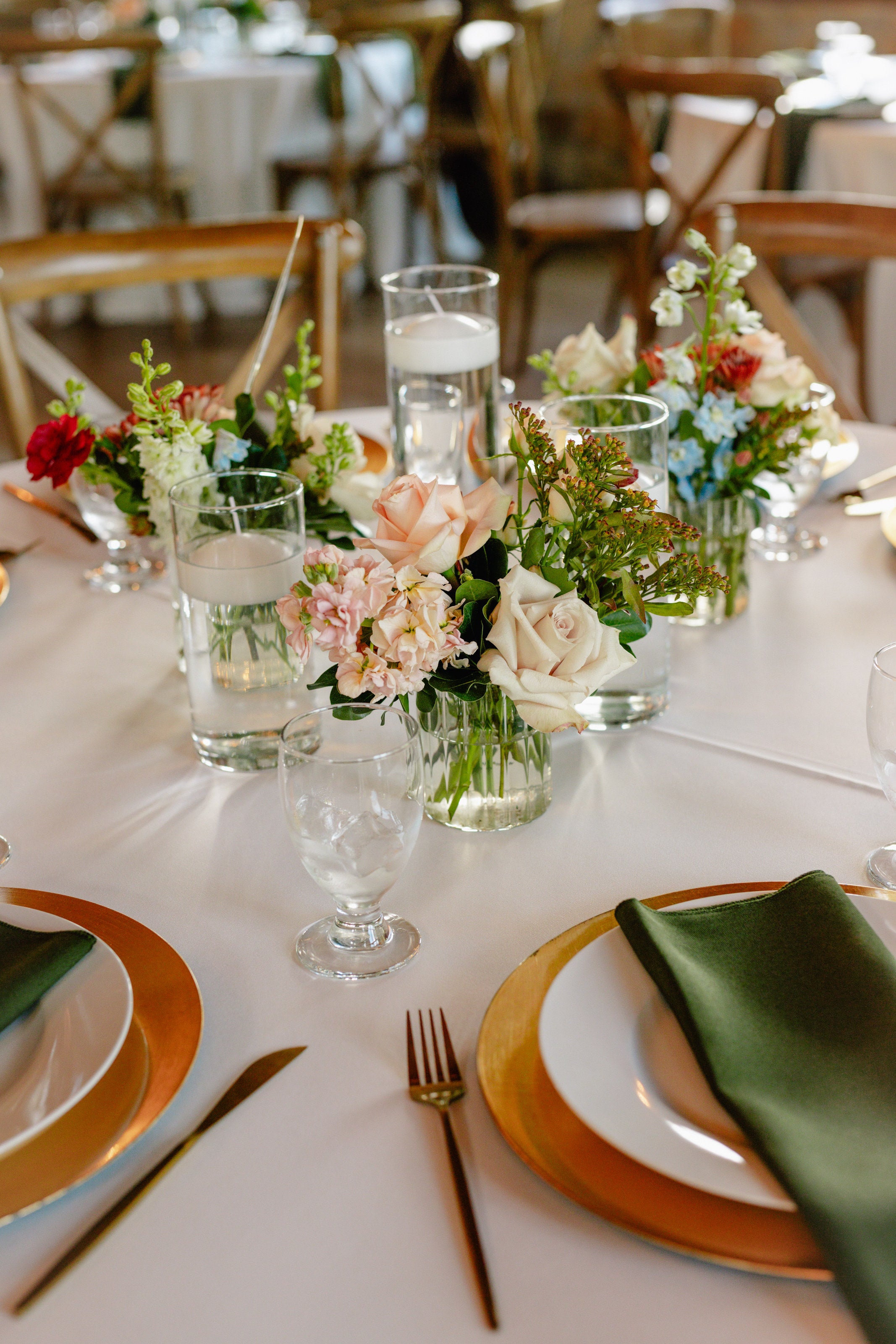 Bud vases clustered down a reception table at Timberview Lodge with cream and peach roses, pale blue delphinium and floating candles, deep olive napkins on gold chargers, by B.blumen