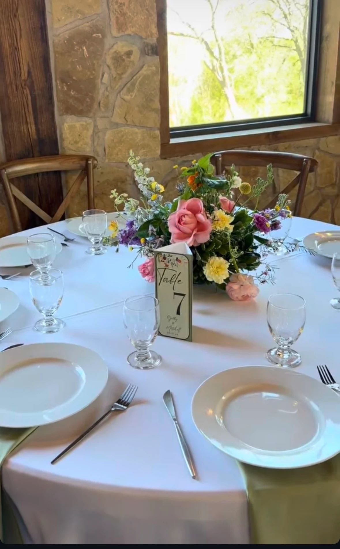 Round reception table at Timberview Lodge with a vibrant wildflower centerpiece of pink garden roses, marigolds, yellow craspedia, and purple delphinium, lime green napkins, against the lodge’s stone wall and window, by B.blumen