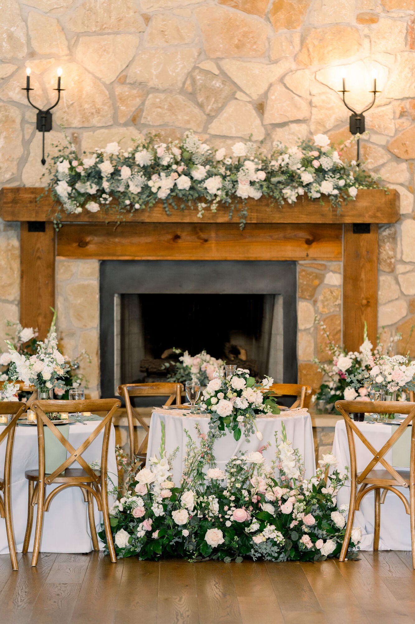 Sweetheart table at Timberview Lodge set against the stone fireplace, with a lush mantel garland of white florals and a cascading ground arrangement at the table’s base, by B.blumen
