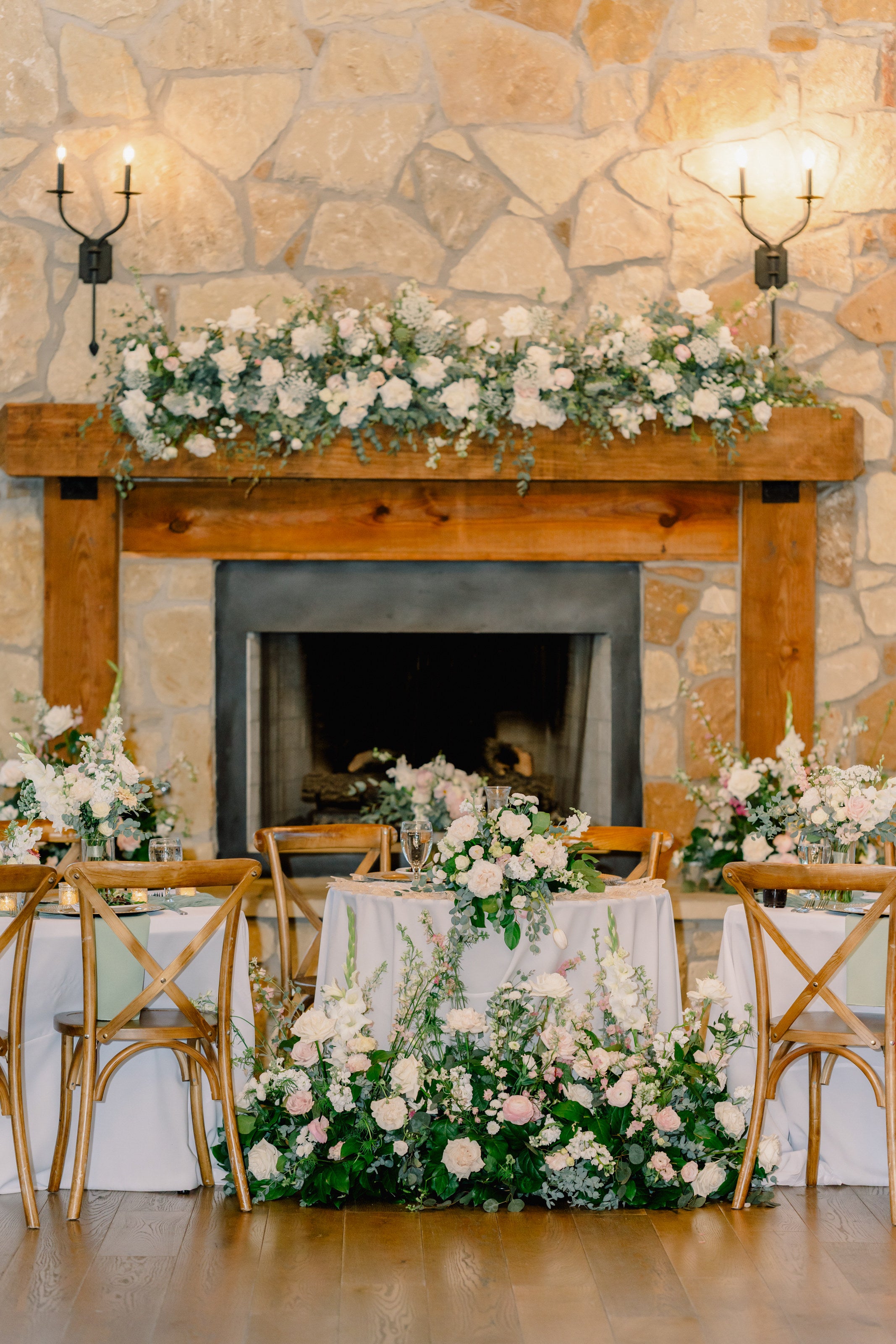 Sweetheart table at Timberview Lodge set against the stone fireplace, with a lush mantel garland of white florals and a cascading ground arrangement at the table’s base, by B.blumen