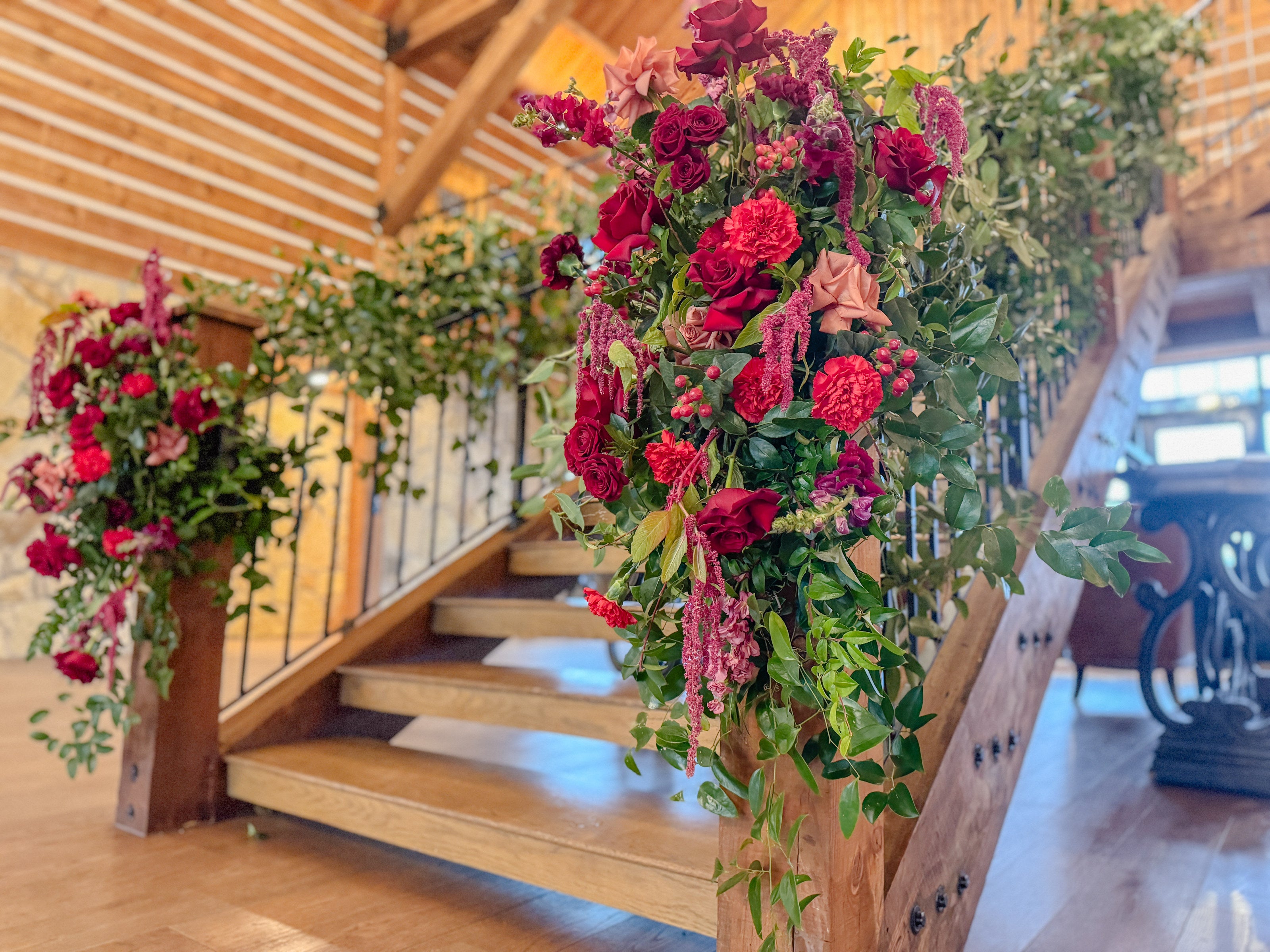 Sweeping floor arrangements of deep red roses, burgundy carnations, blush garden roses, and hanging amaranthus flanking the wood-and-iron staircase at Timberview Lodge, by B.blumen