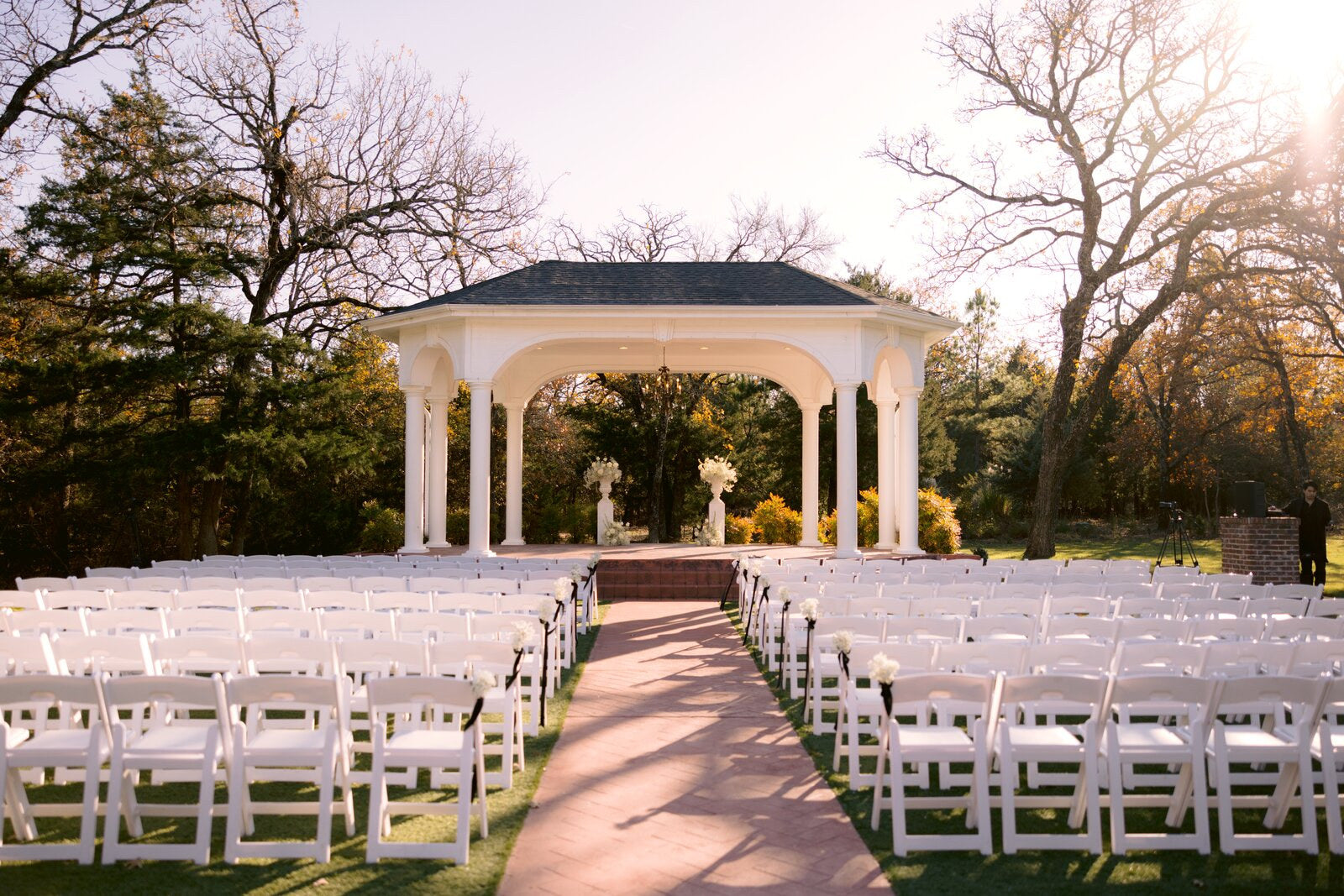 Outdoor ceremony at Rockwall Manor under the white-columned classical pavilion, brick aisle, rows of white garden chairs, twin floral arrangements at the altar, by B.blumen