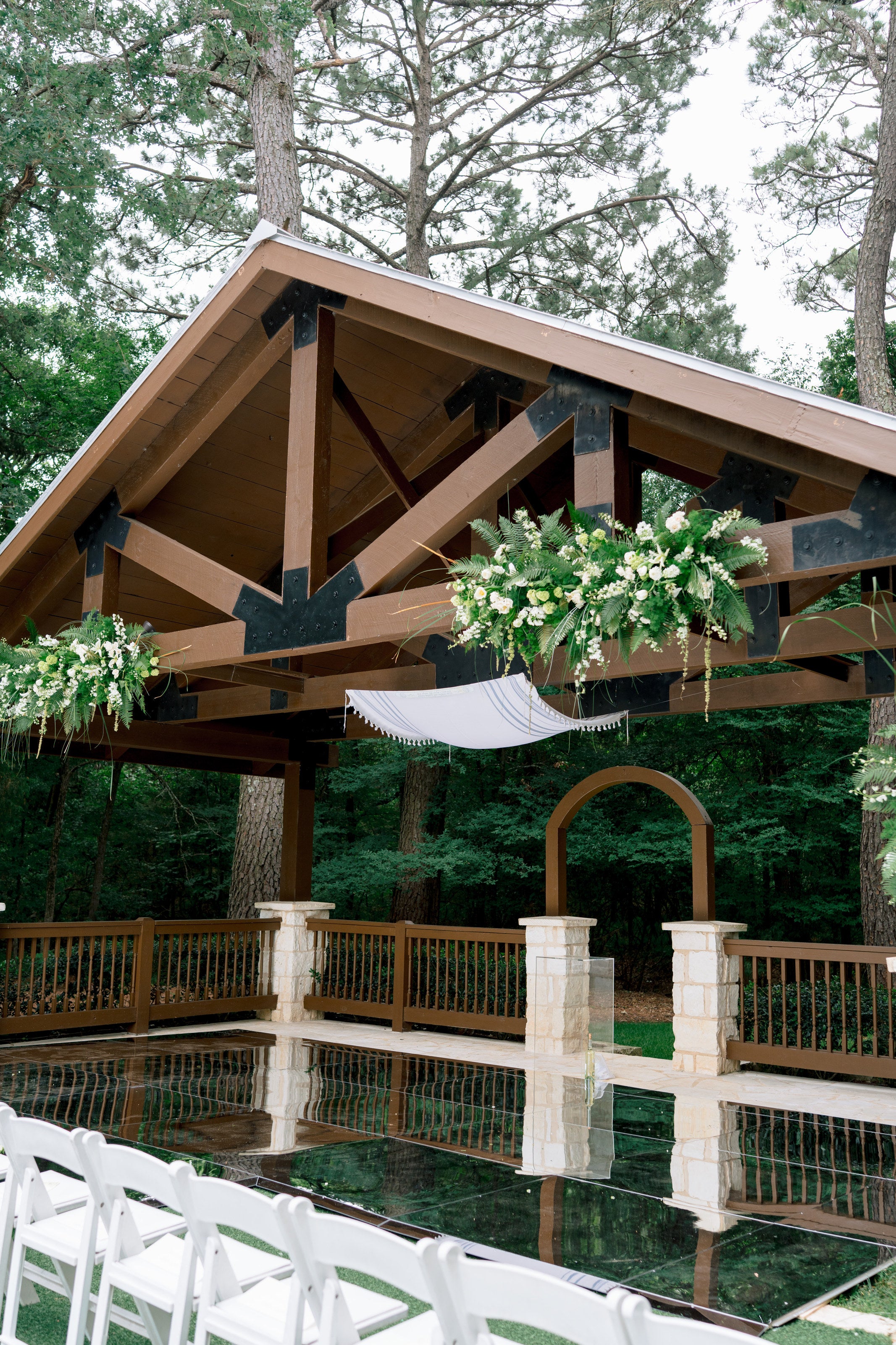 The wood pavilion at Poetry Springs, greenery and white florals mounted along the timber trusses over a canvas sling, a stone arch behind and white chairs at the reflecting pool, by B.blumen