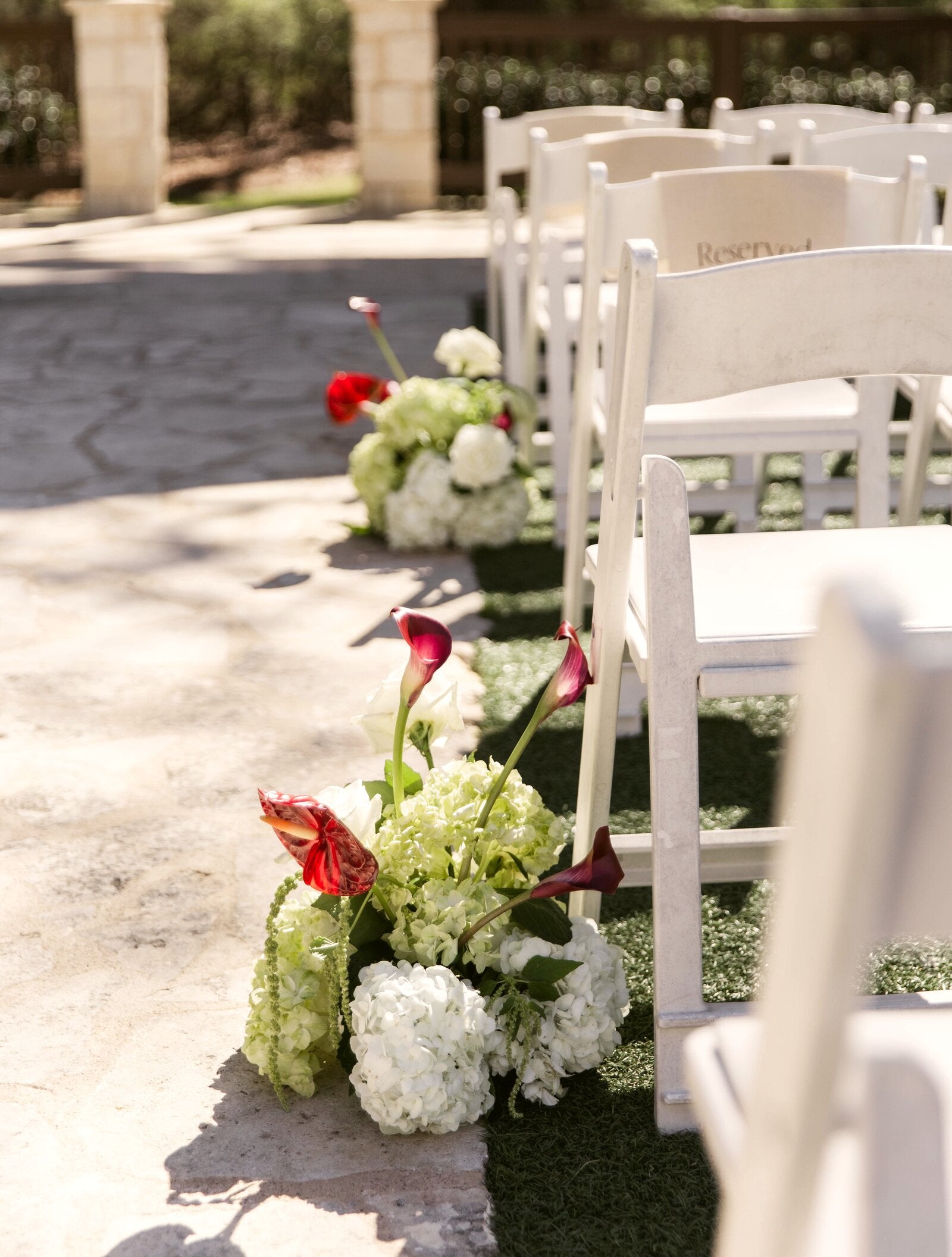 Outdoor ceremony aisle on a stone path at The Springs Rockwall with reserved white garden chairs and aisle markers of deep red calla lilies, hydrangea, and trailing greenery, by B.blumen