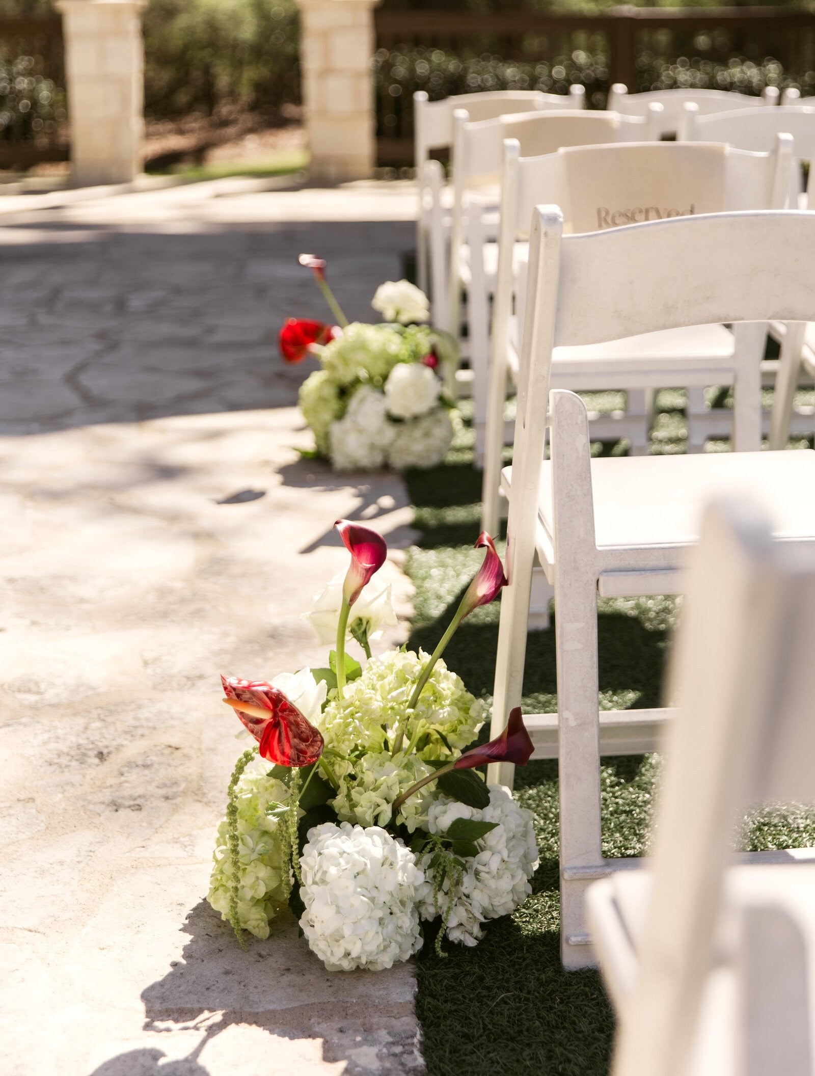 Outdoor ceremony aisle on a stone path at The Springs Rockwall with reserved white garden chairs and aisle markers of deep red calla lilies, hydrangea, and trailing greenery, by B.blumen