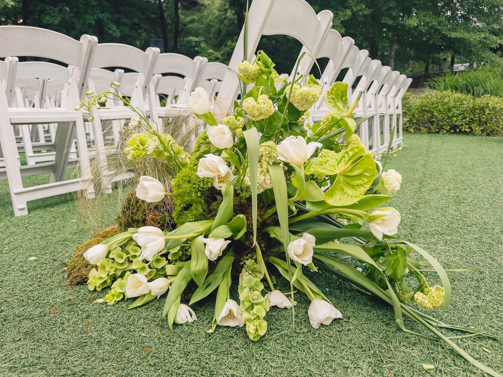 Aisle marker at Poetry Springs with white tulips, callas, ranunculus, hydrangea, bells of Ireland, and trailing greenery on a moss base, set on green turf in front of white garden chairs, by B.blumen