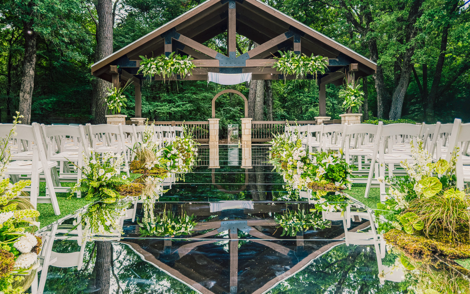 Outdoor wood-beam pavilion ceremony at Poetry Springs with a mirror aisle running down the center, a white fabric drape suspended under the peak, lush white and green florals at each row, white garden chairs flanking, by B.blumen
