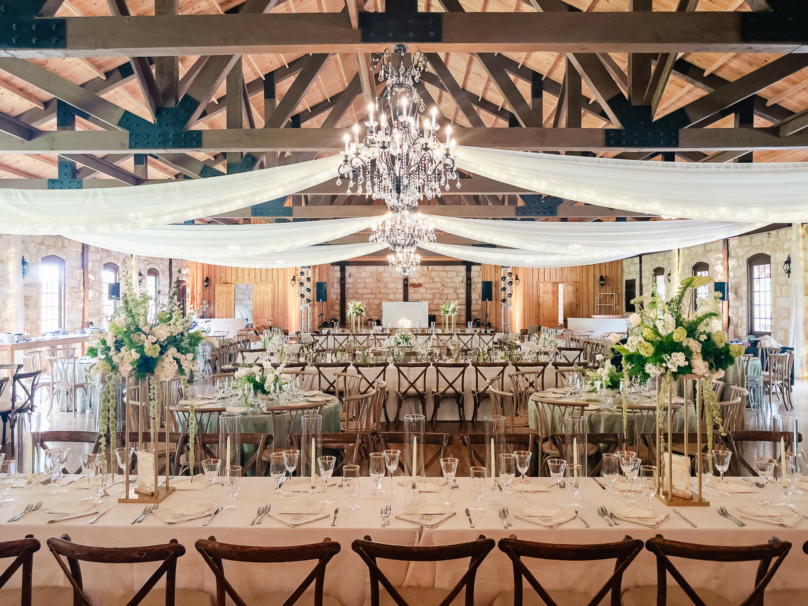 Wide reception establishing at Poetry Springs with the dark exposed truss ceiling draped in white fabric and twinkle lights, crystal chandelier center, head table in the distance, round tables with tall pedestal florals in the foreground, by B.blumen