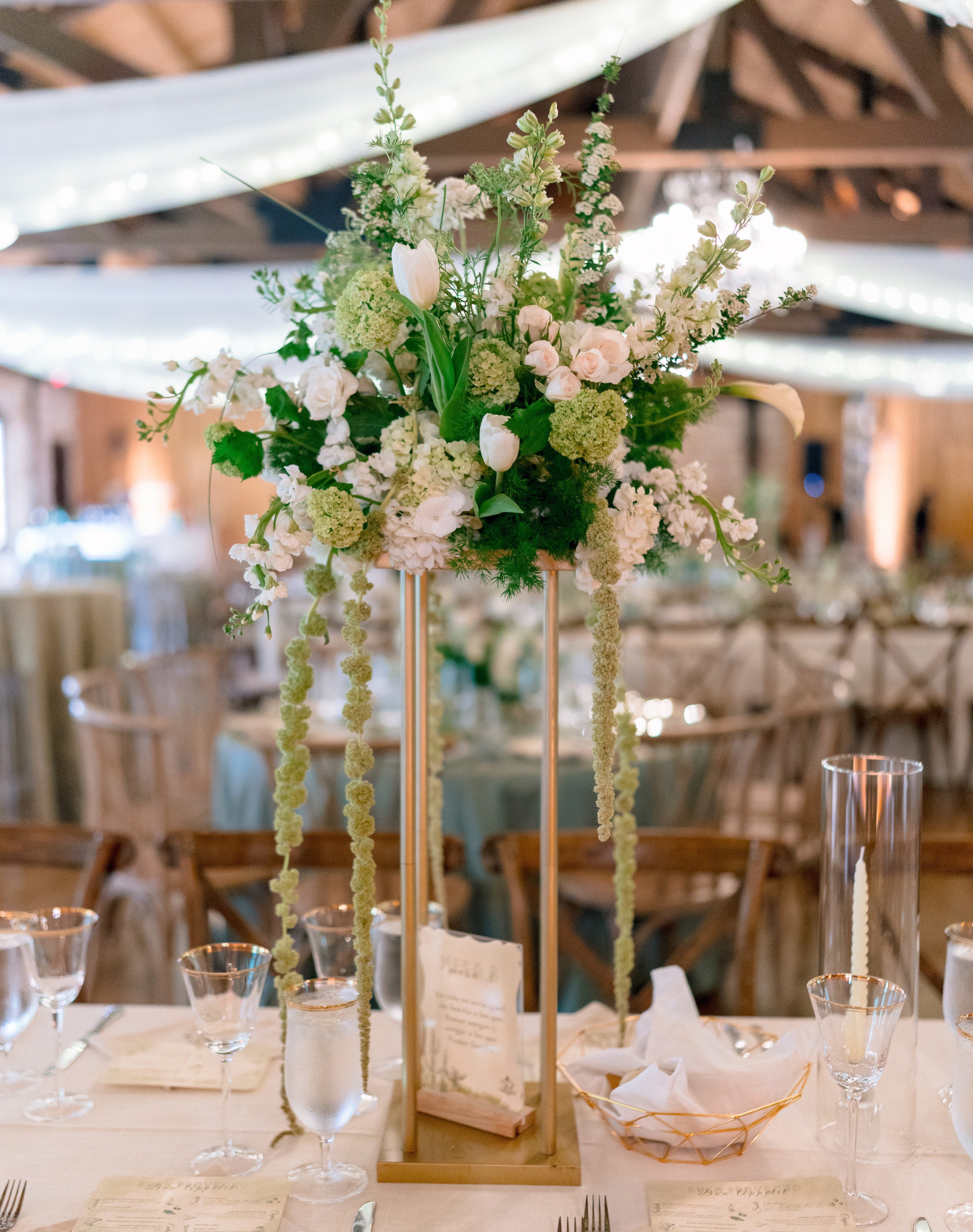 Tall gold-stand centerpiece at The Springs Rockwall in white roses, stock and green foliage, above a set table under draped ceiling fabric, by B.blumen