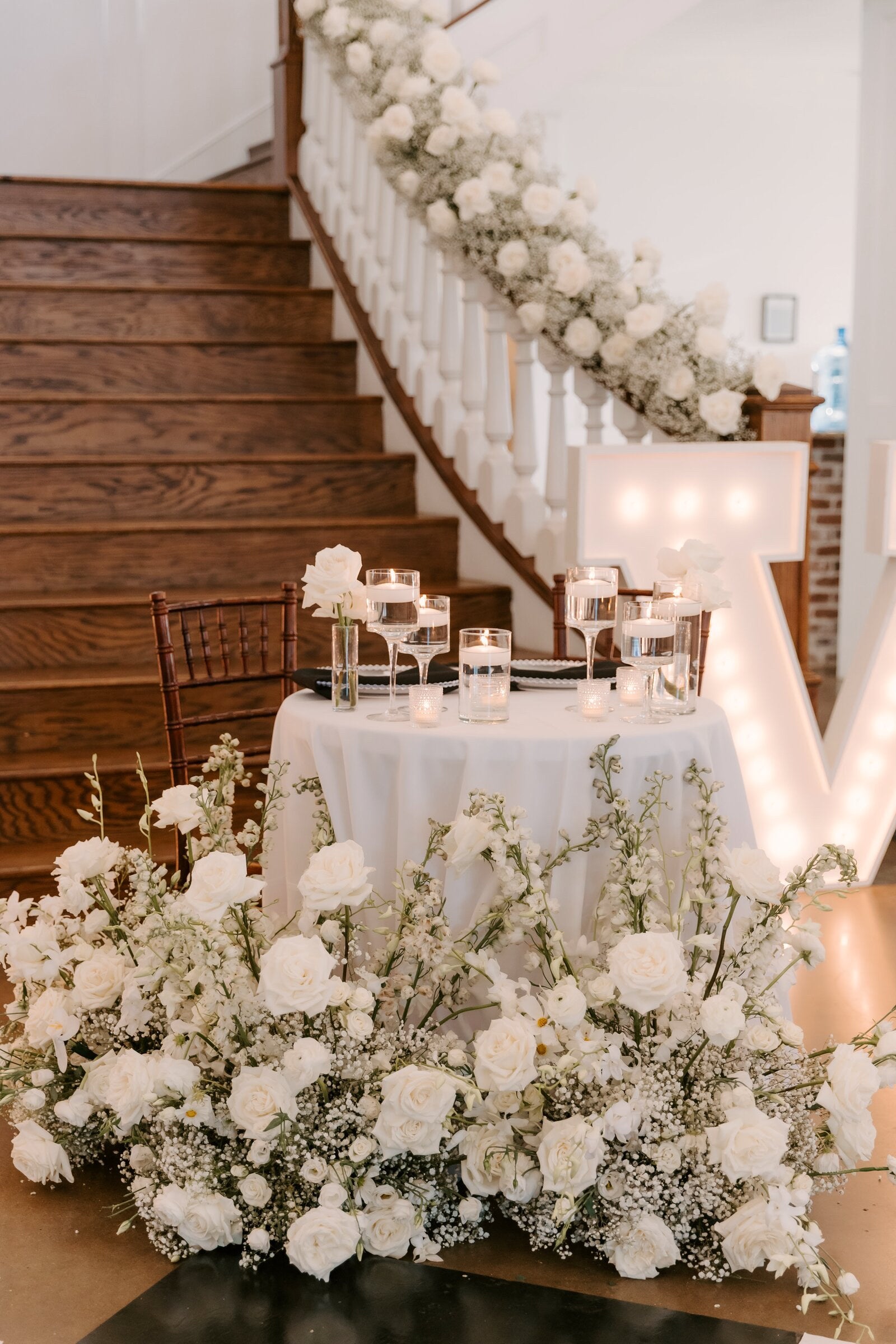 Sweetheart table at the base of the Rockwall Manor staircase with a flowing baby's breath garland down the banister, a marquee letter at the side, and a lush white ground arrangement at the table's base, by B.blumen