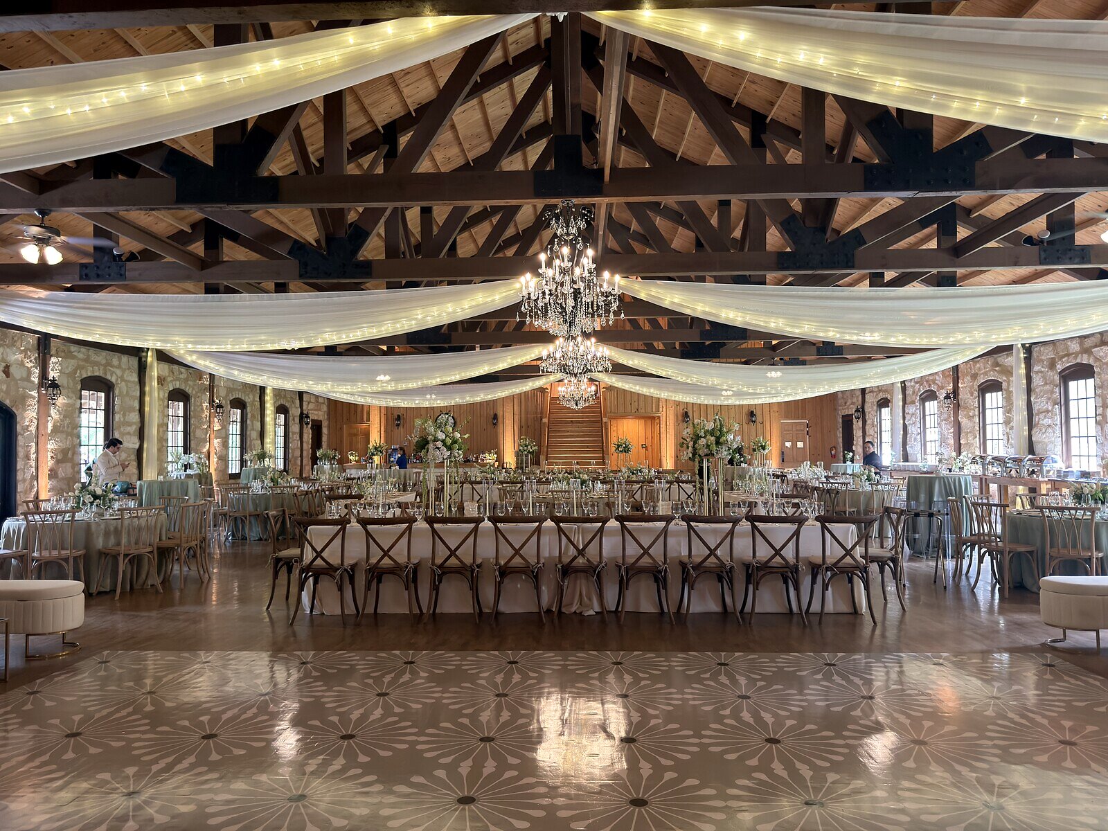 Widest reception view at Poetry Springs with the mandala-pattern dance floor in the foreground, head table on bench seating in the distance, two crystal chandeliers, and draped ceiling fabric with twinkle lights, by B.blumen