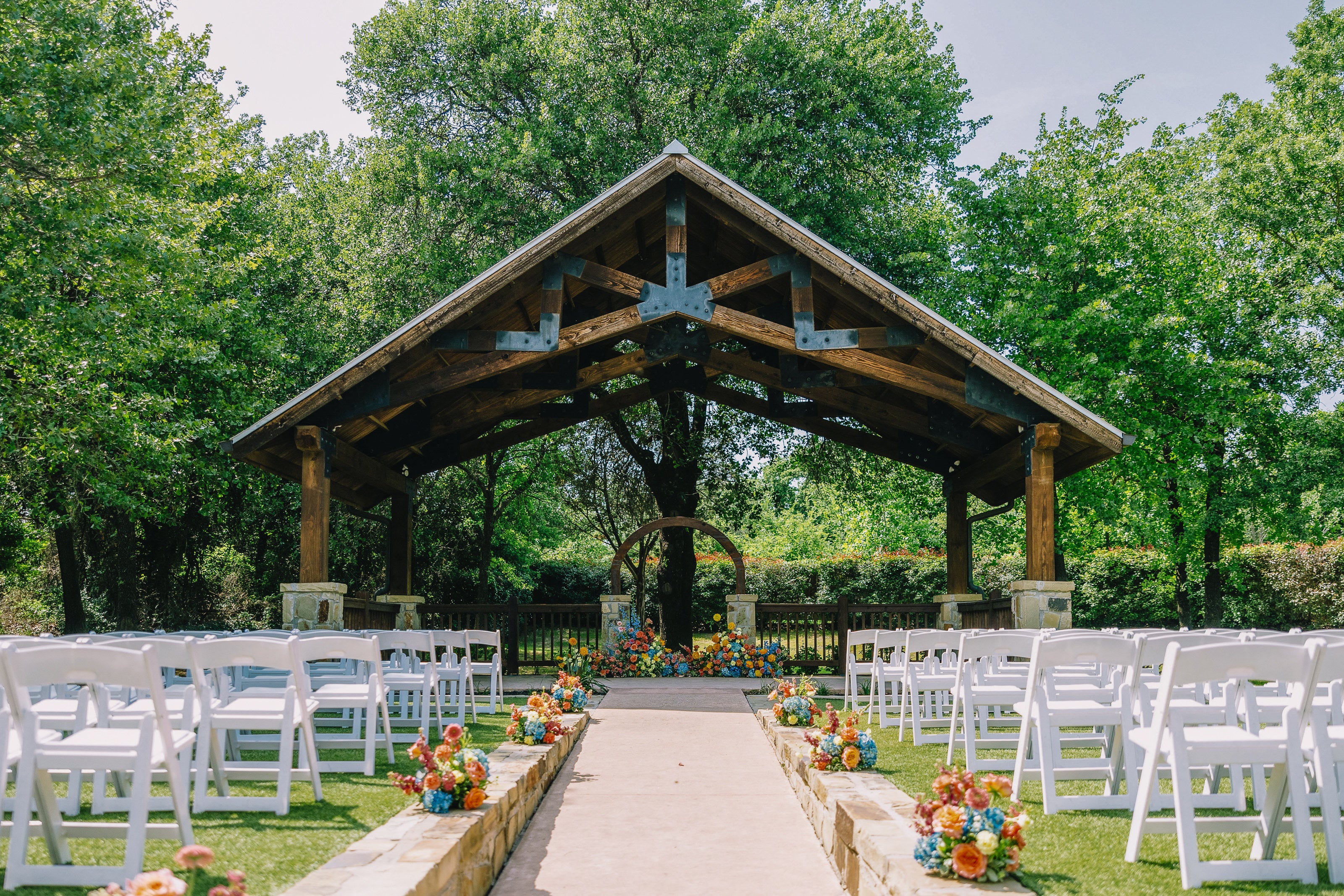 Wood pavilion outdoor ceremony at The Springs Weatherford Westwood Hall, with stone aisle walls and vibrant orange, blue, and yellow florals by B.blumen