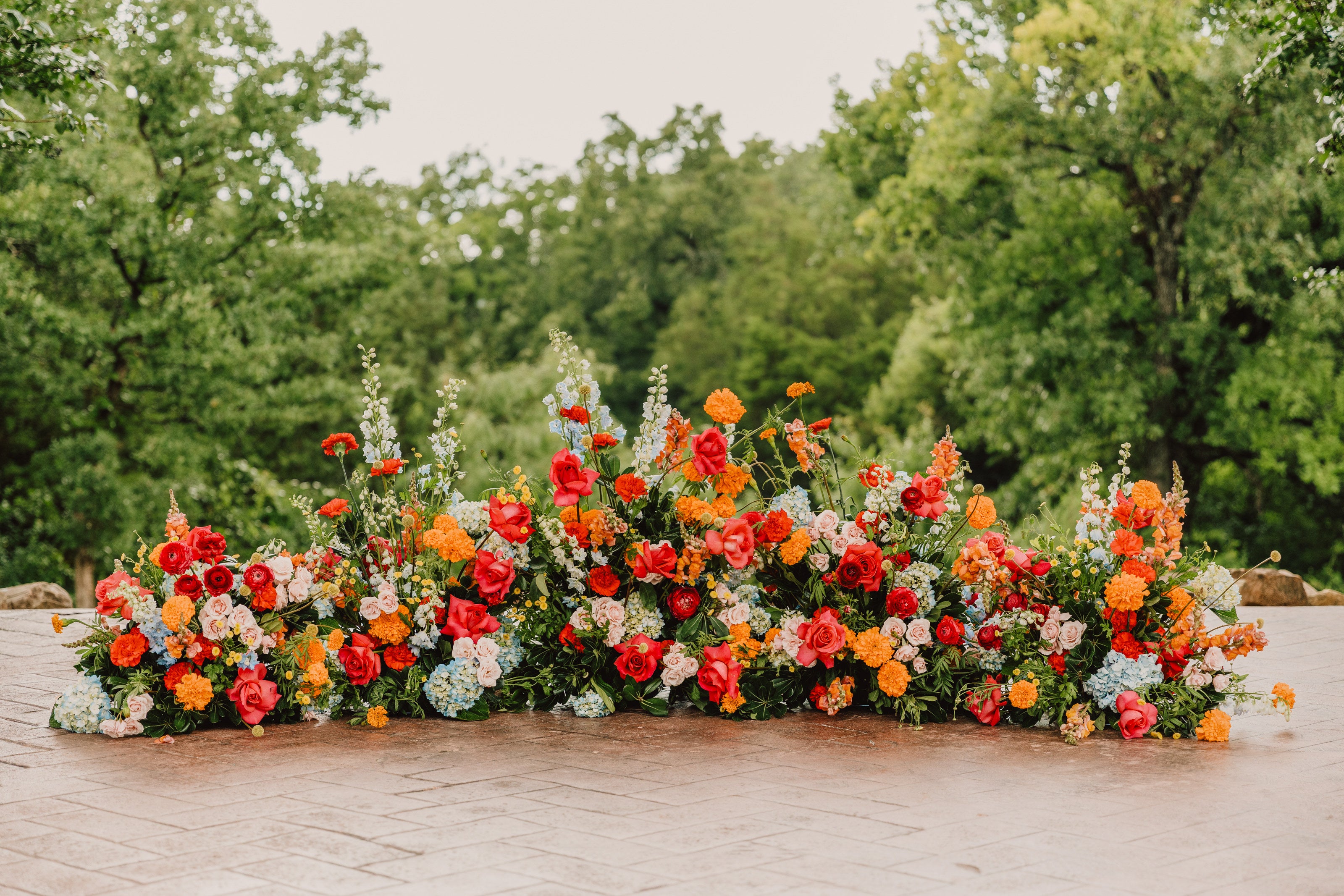 Sweeping ground arrangement of coral, red, blue, and white florals on the brick terrace at Parker Manor, by B.blumen
