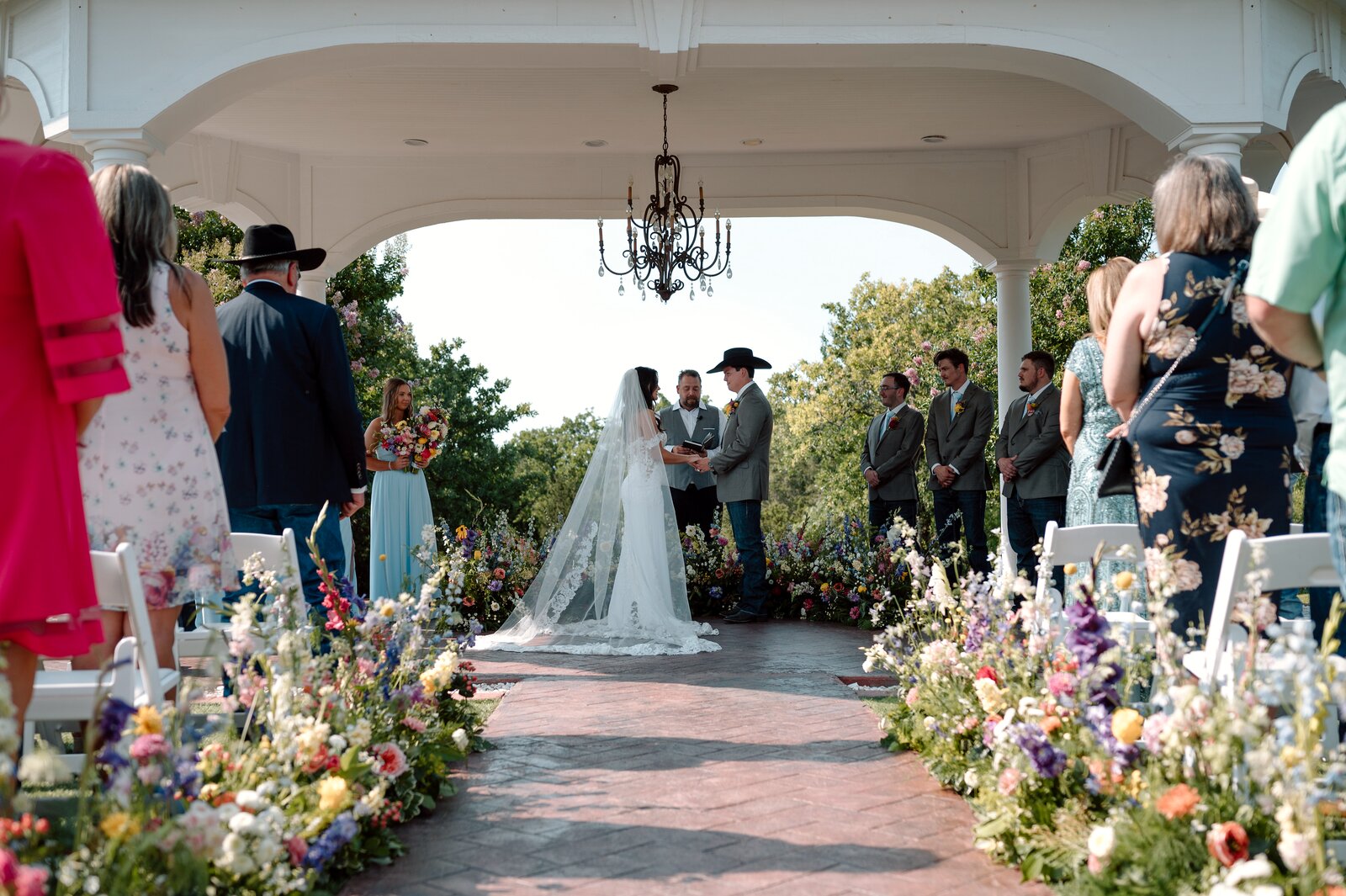 Outdoor pergola ceremony at Parker Manor with brick aisle lined in wildflower markers, brass chandelier above the altar, by B.blumen