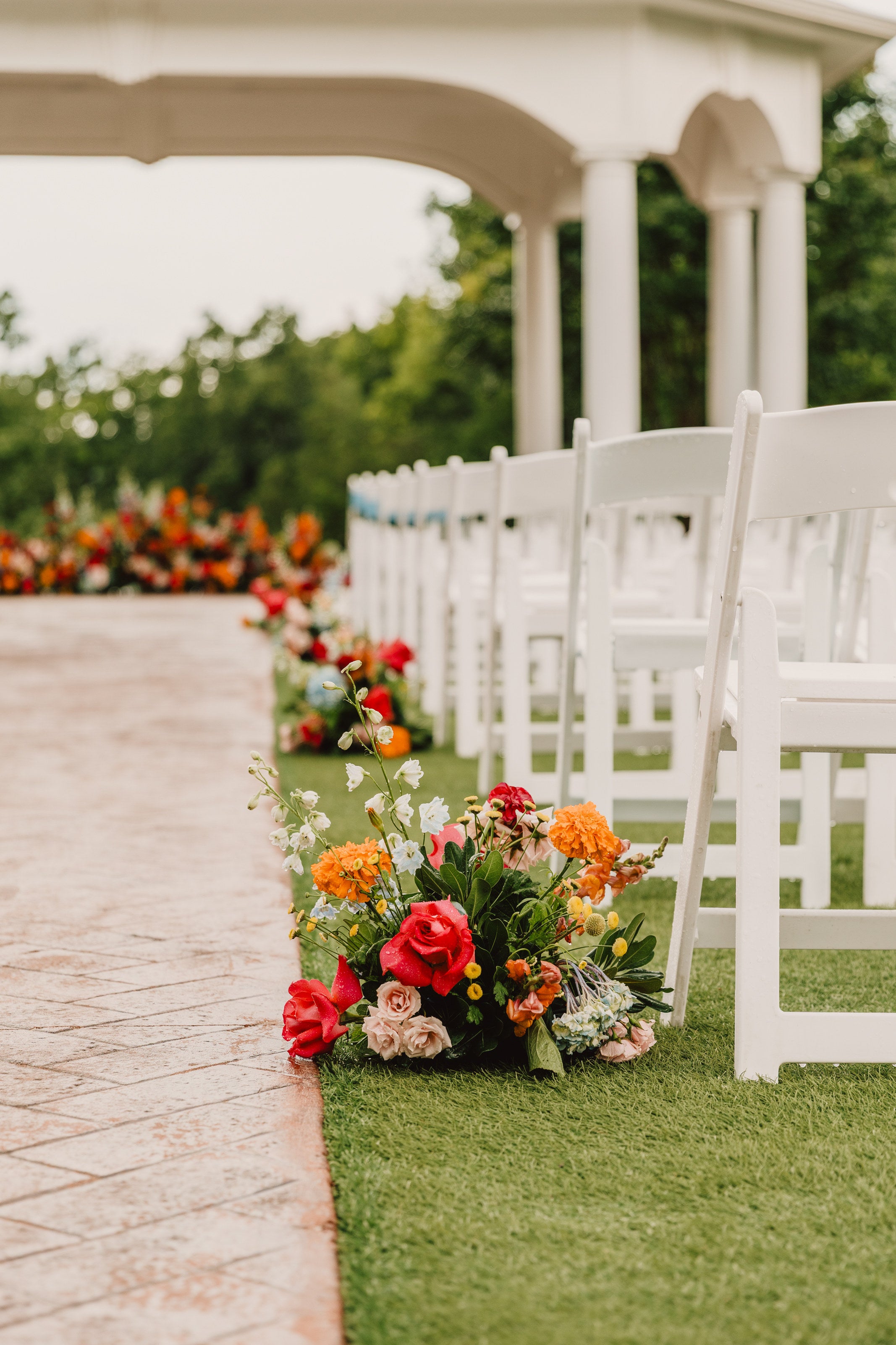 Aisle clusters of orange, coral, and red garden florals along white chairs at the Parker Manor pavilion by B.blumen
