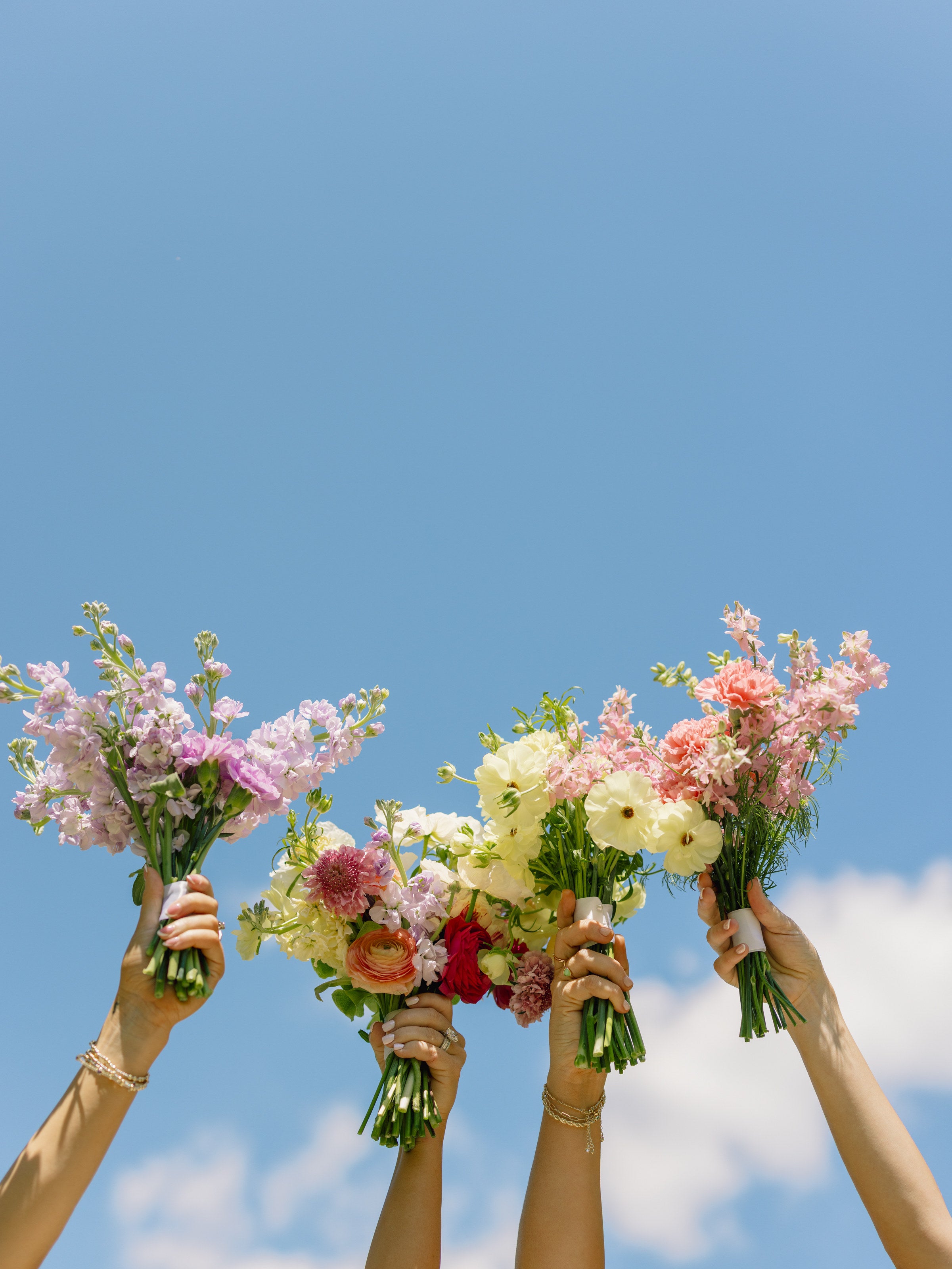 Four bridesmaid bouquets in pink, coral, and white raised against a blue Texas sky at The Springs Weatherford, by B.blumen
