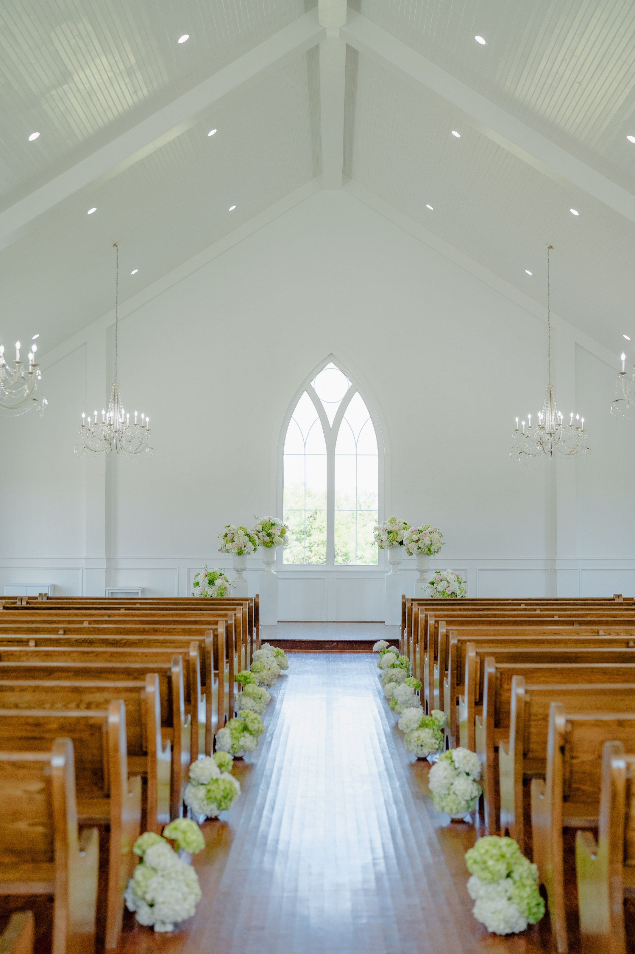 Interior of The Springs Weatherford chapel with wood pews, white walls, and white-and-green pew florals leading to the altar, by B.blumen