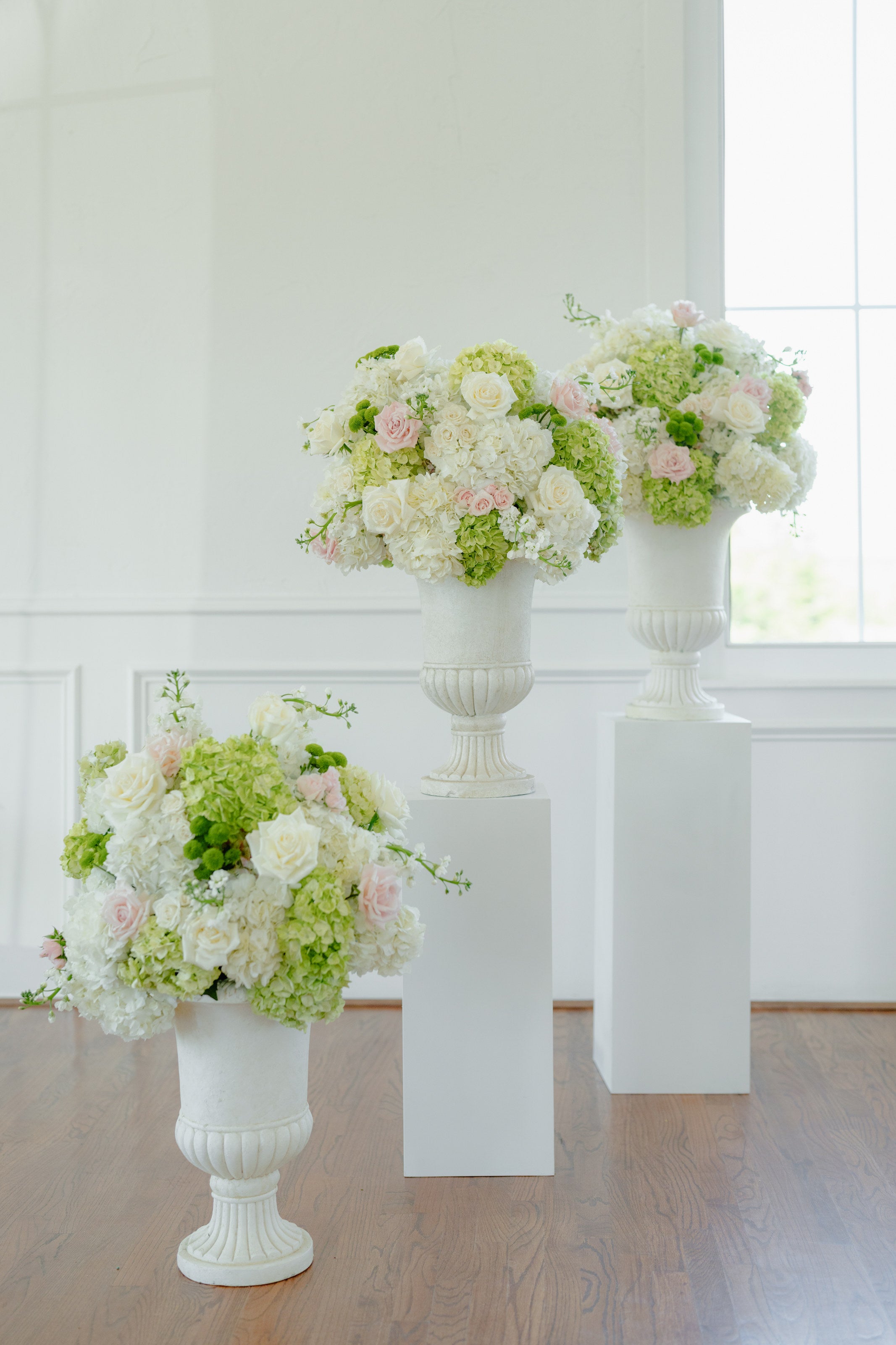 White urn arrangements of hydrangea, garden roses, and viburnum on plinths beside the arched window of The Springs Weatherford chapel, by B.blumen