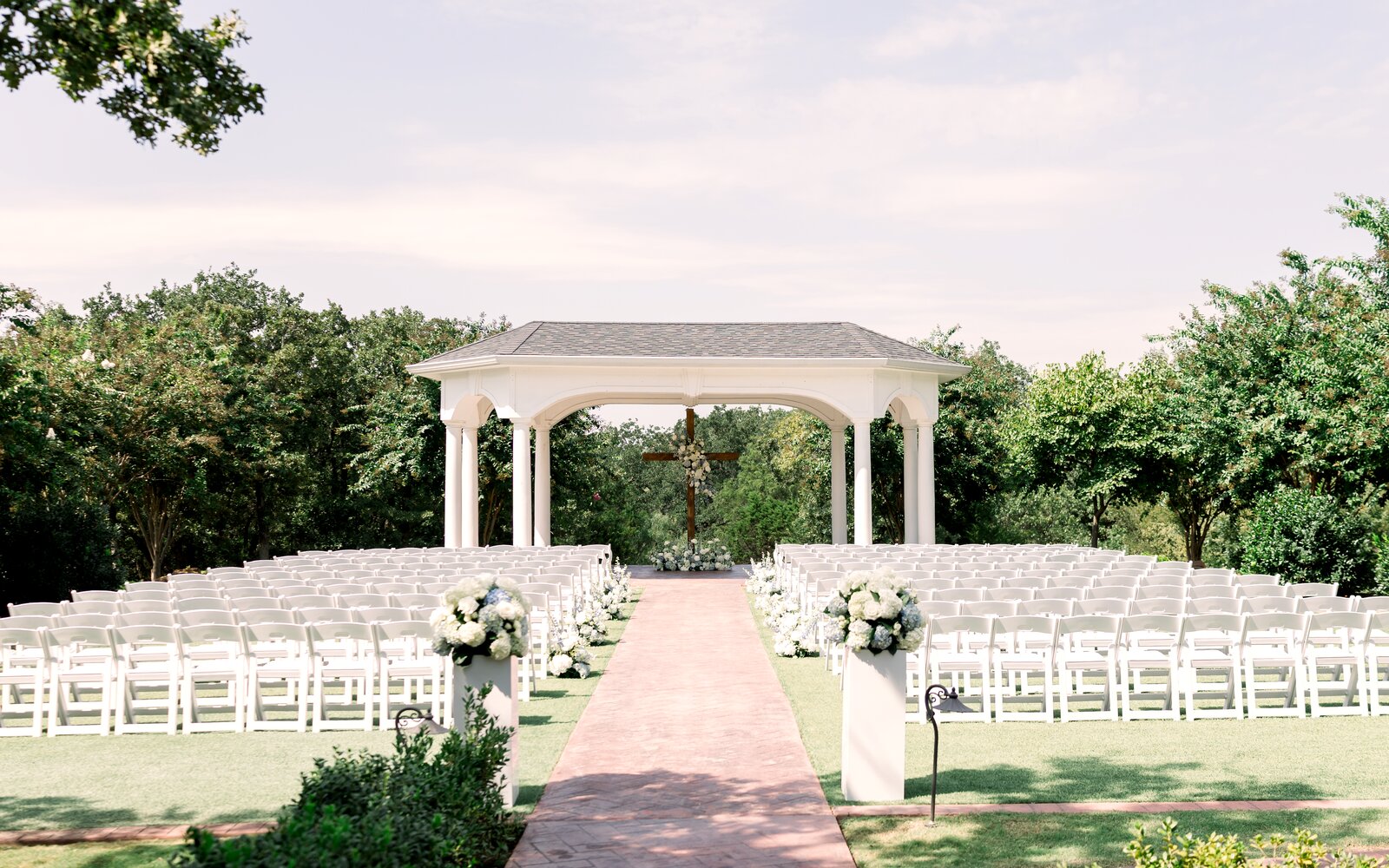 Outdoor ceremony at The Springs Weatherford under the white pergola, brick aisle lined with white pedestal florals by B.blumen