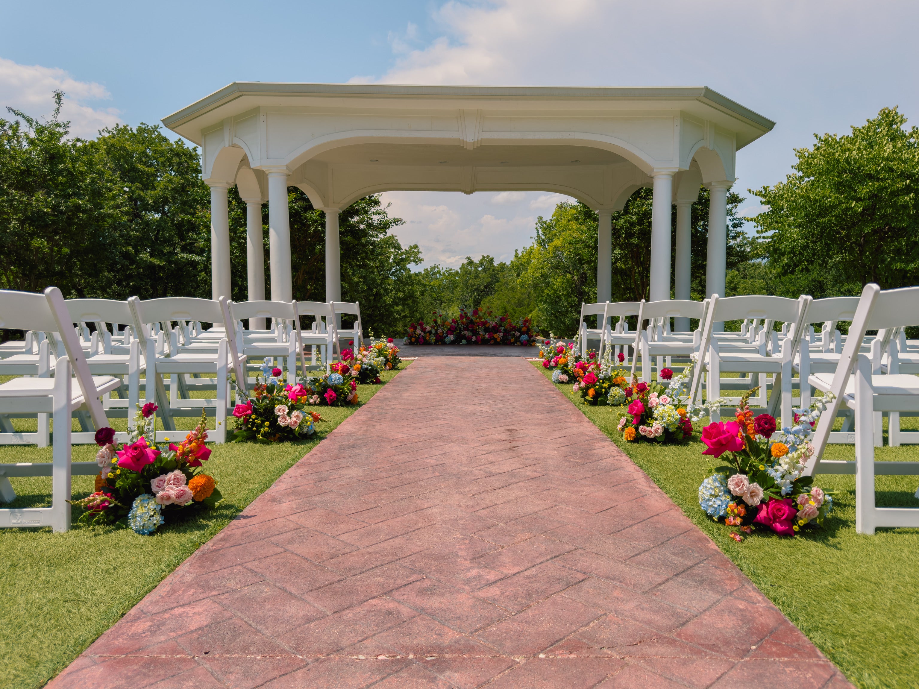 Outdoor ceremony at The Springs Weatherford, white gazebo at the head of a brick aisle lined with vivid pink, orange and blue florals and rows of white garden chairs, by B.blumen