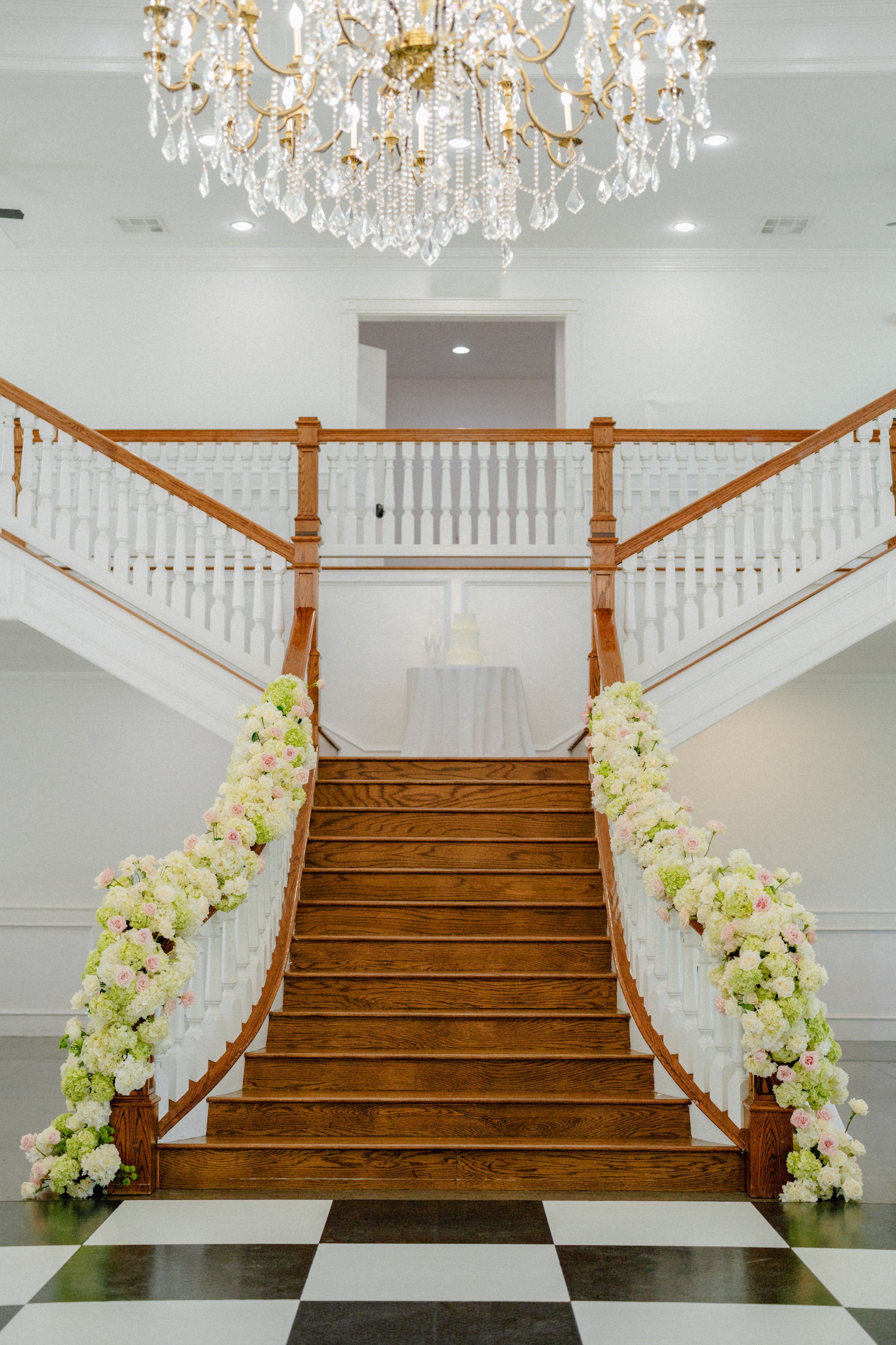 Grand staircase at Parker Manor wrapped in white hydrangea garland over the checkered floor, chandelier above, by B.blumen