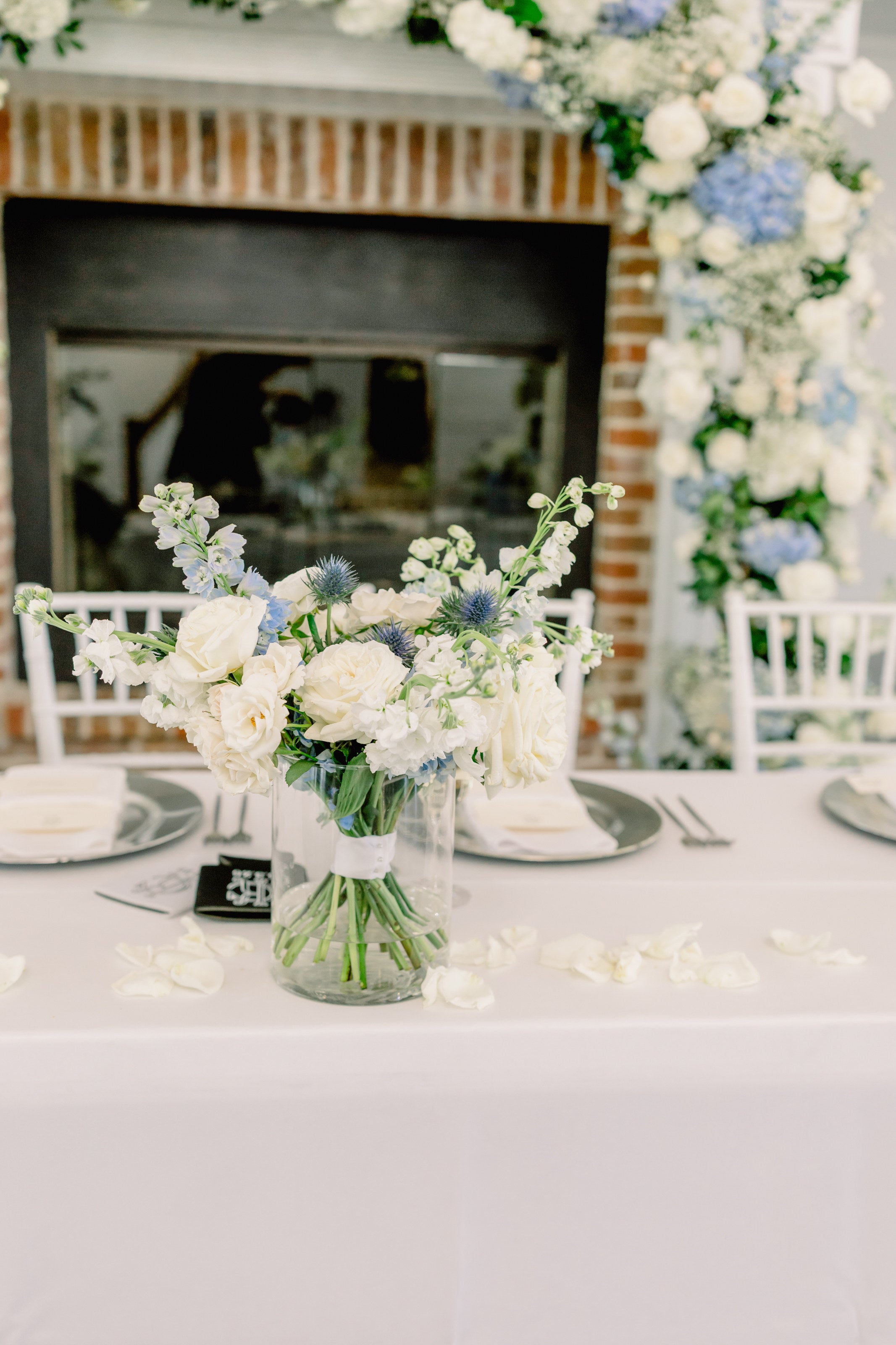 Reception table at Parker Manor with blue and white florals in glass, set before the brick fireplace, by B.blumen
