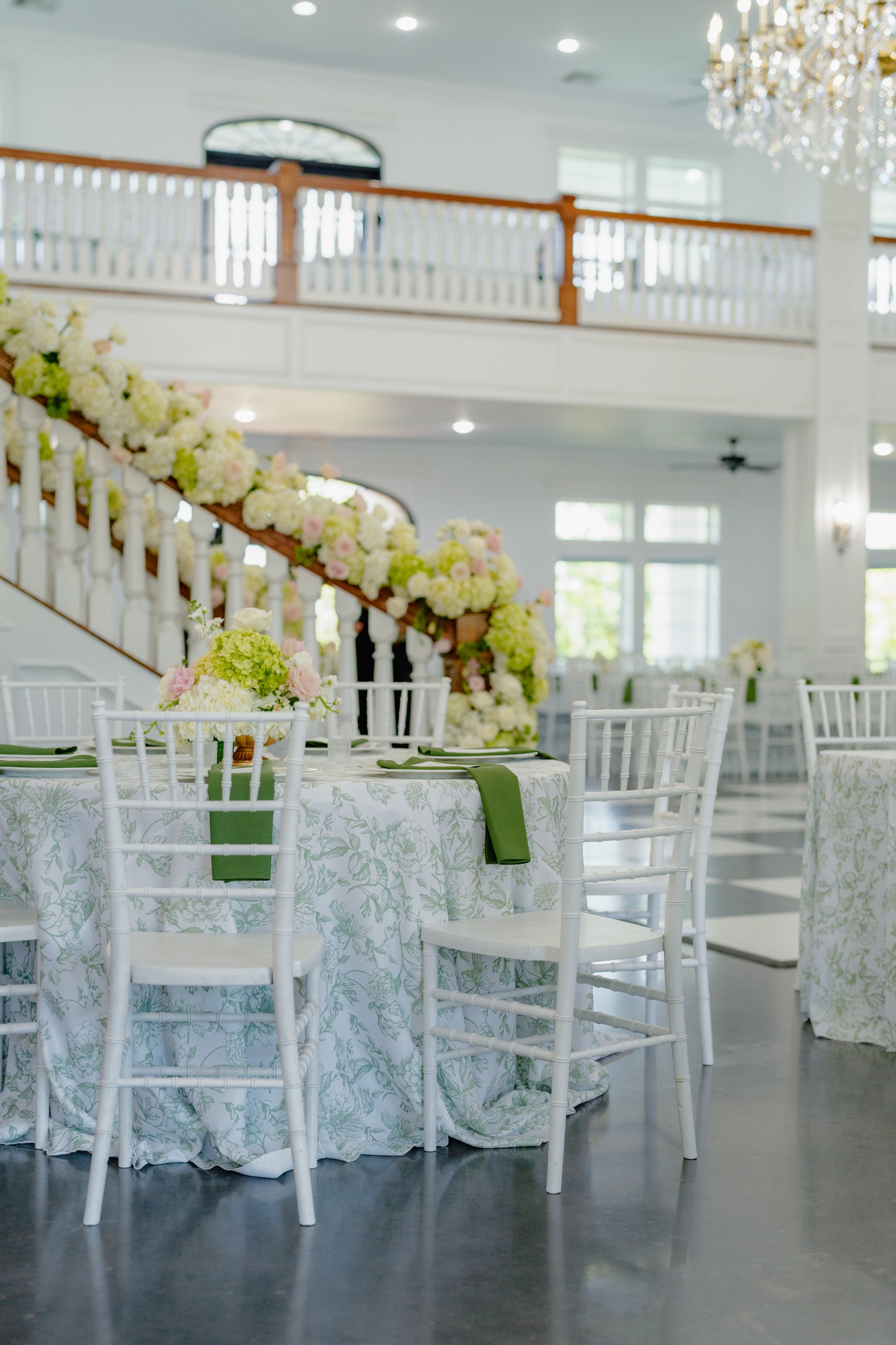 Reception tables at Parker Manor in pale blue lace linens with white floral centerpieces, the garlanded staircase behind, by B.blumen