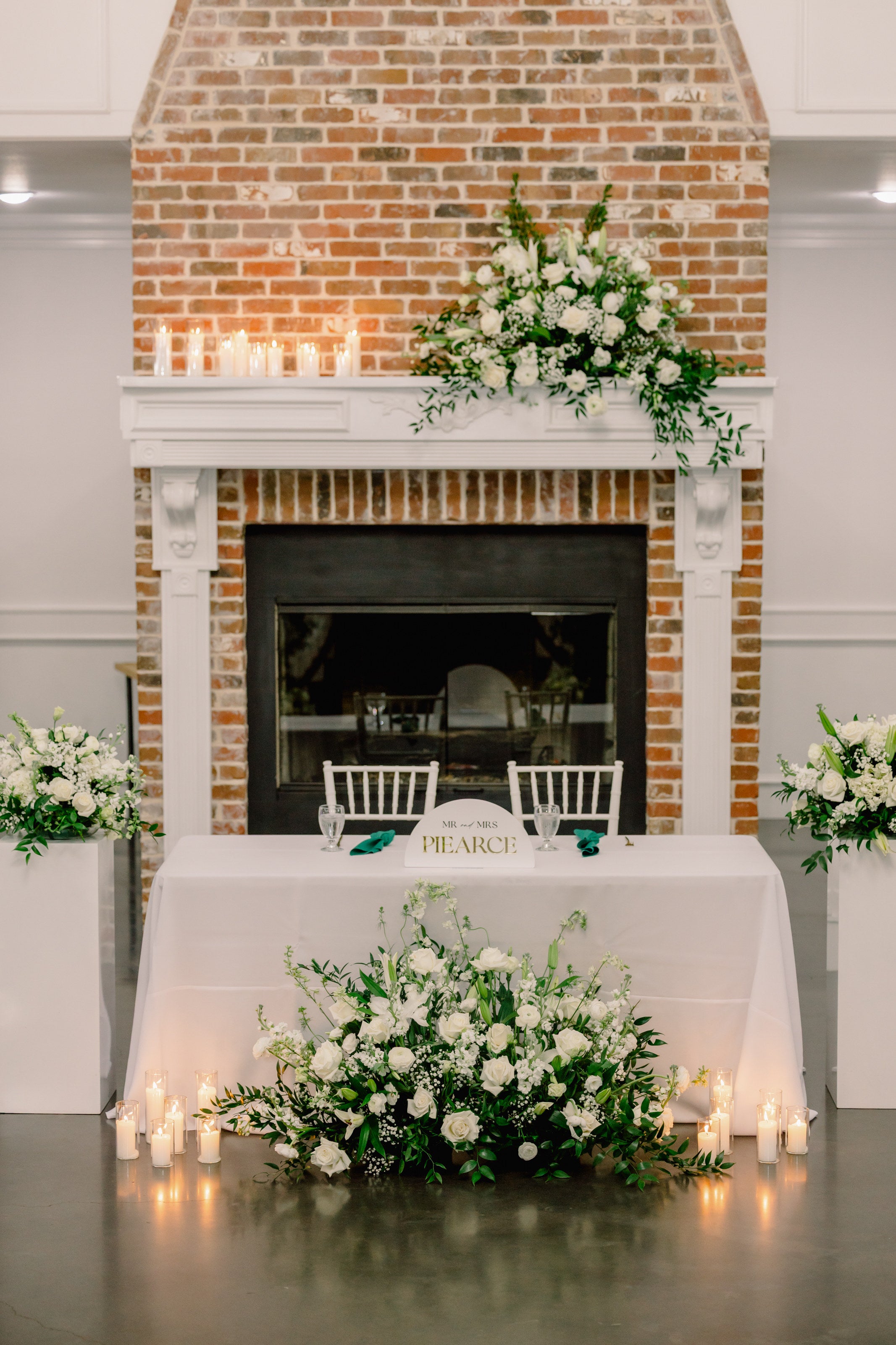 Sweetheart table at Parker Manor set against a brick fireplace, with white florals on the mantle and a candlelit ground arrangement at its base by B.blumen