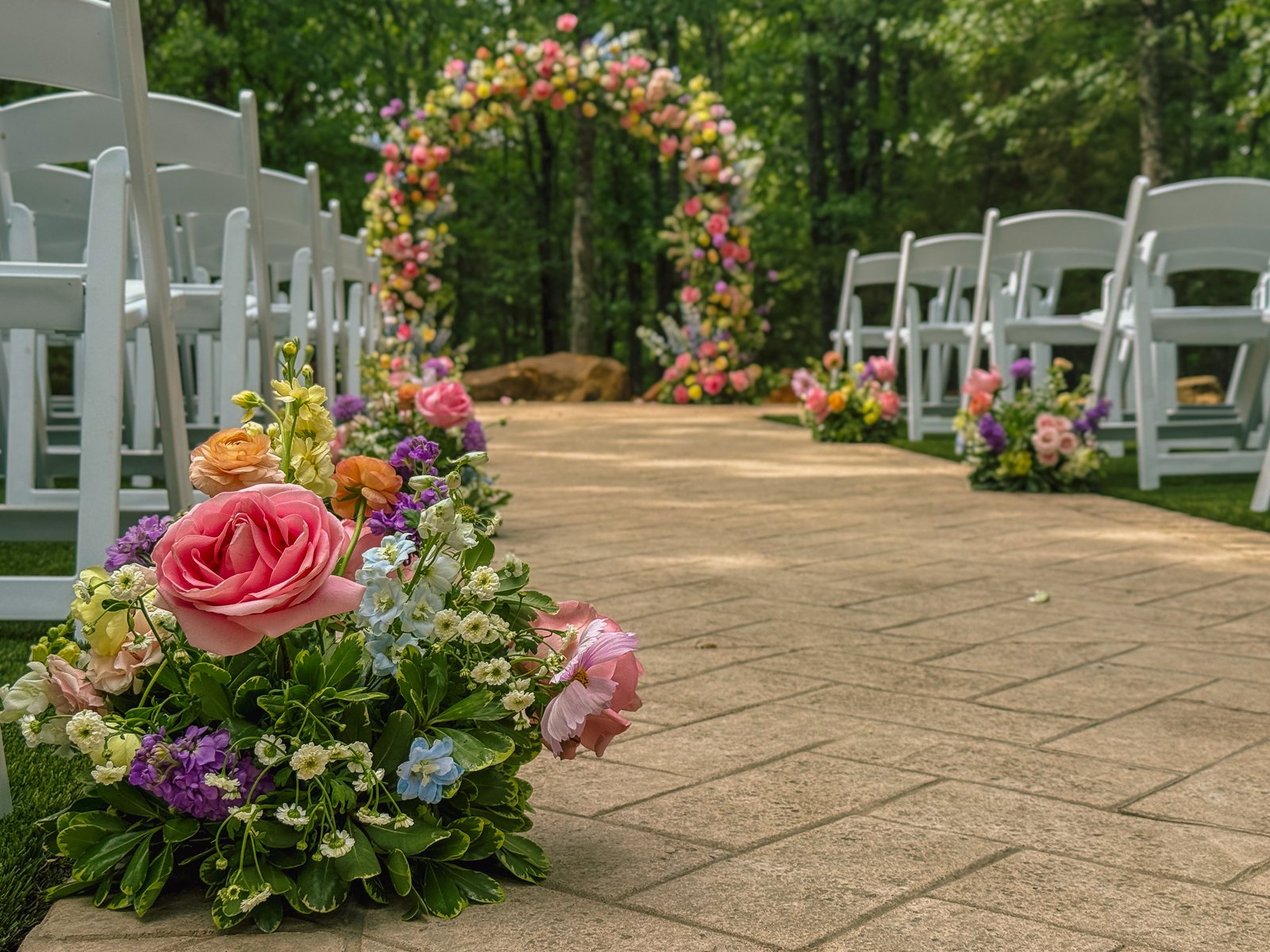 Low-angle view up the ceremony aisle at The White Sparrow, a pink garden-rose aisle arrangement in the foreground and the pastel floral arch beyond, by B.blumen