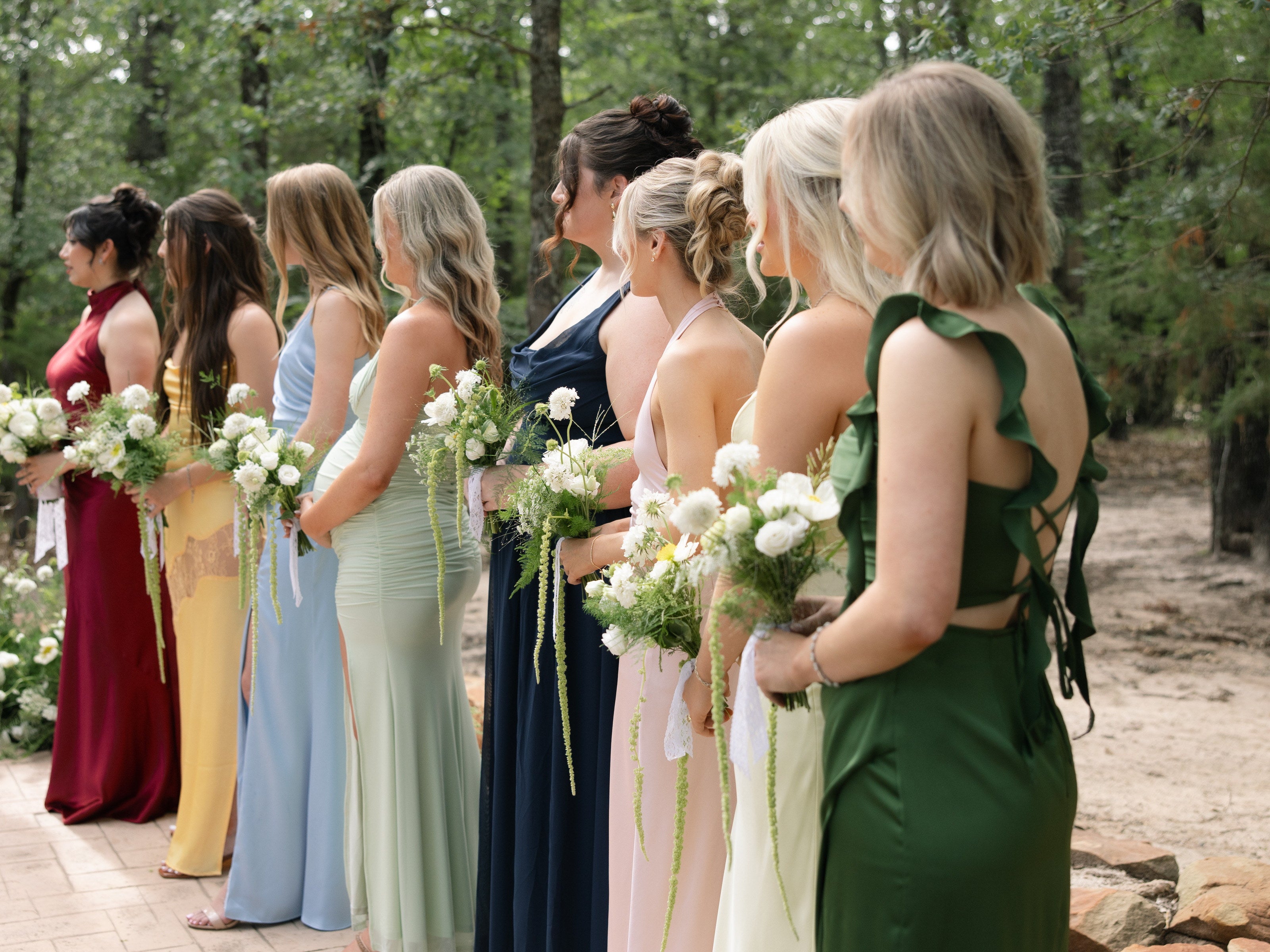 Bridal party standing along the paved ceremony aisle at The White Sparrow in mismatched jewel and pastel gowns, each holding a white and green bouquet with trailing amaranthus, by B.blumen