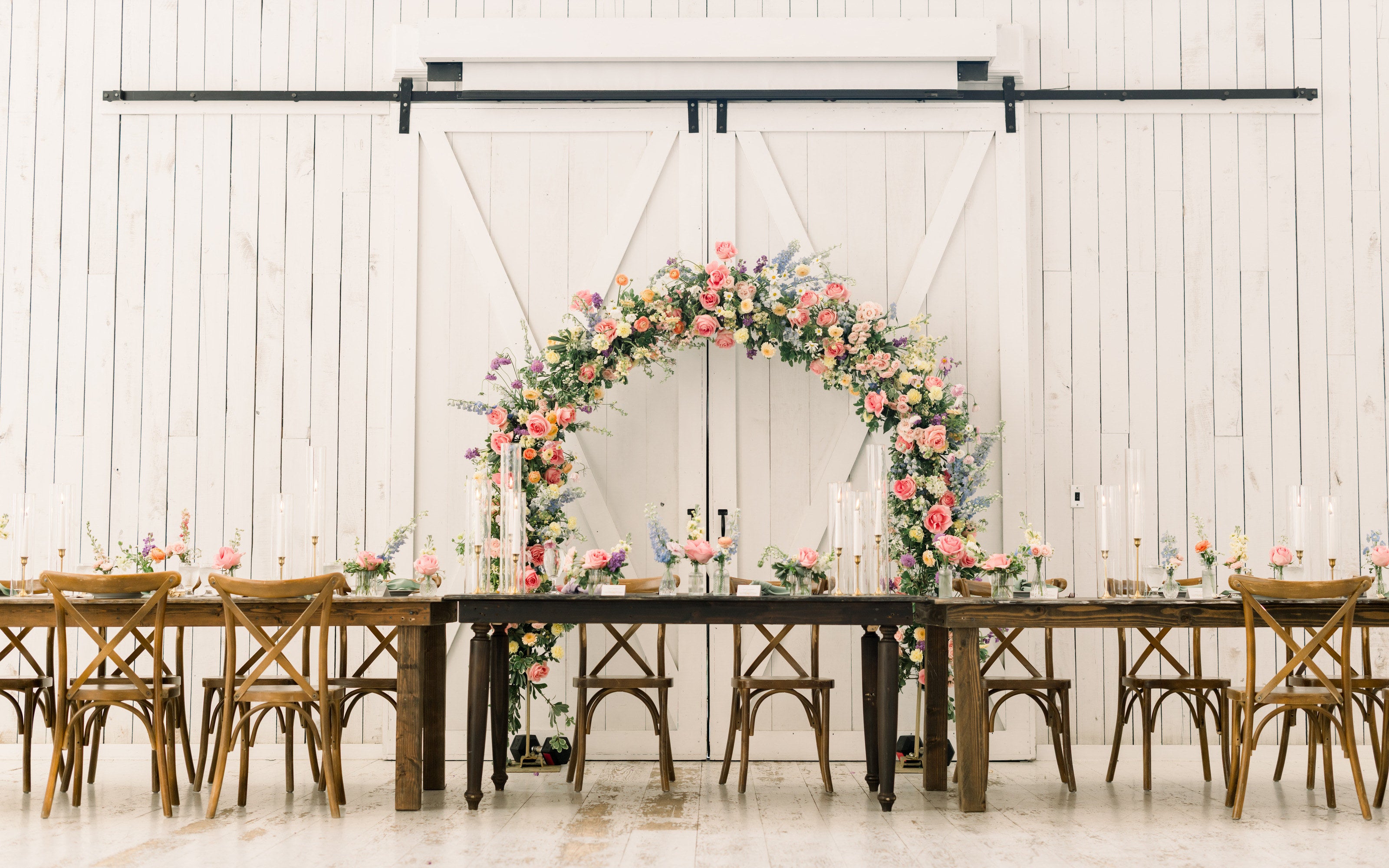 Outdoor ceremony arch at The White Sparrow set against the barn’s white sliding doors, pink and green florals over the paved aisle with cross-back chairs, by B.blumen