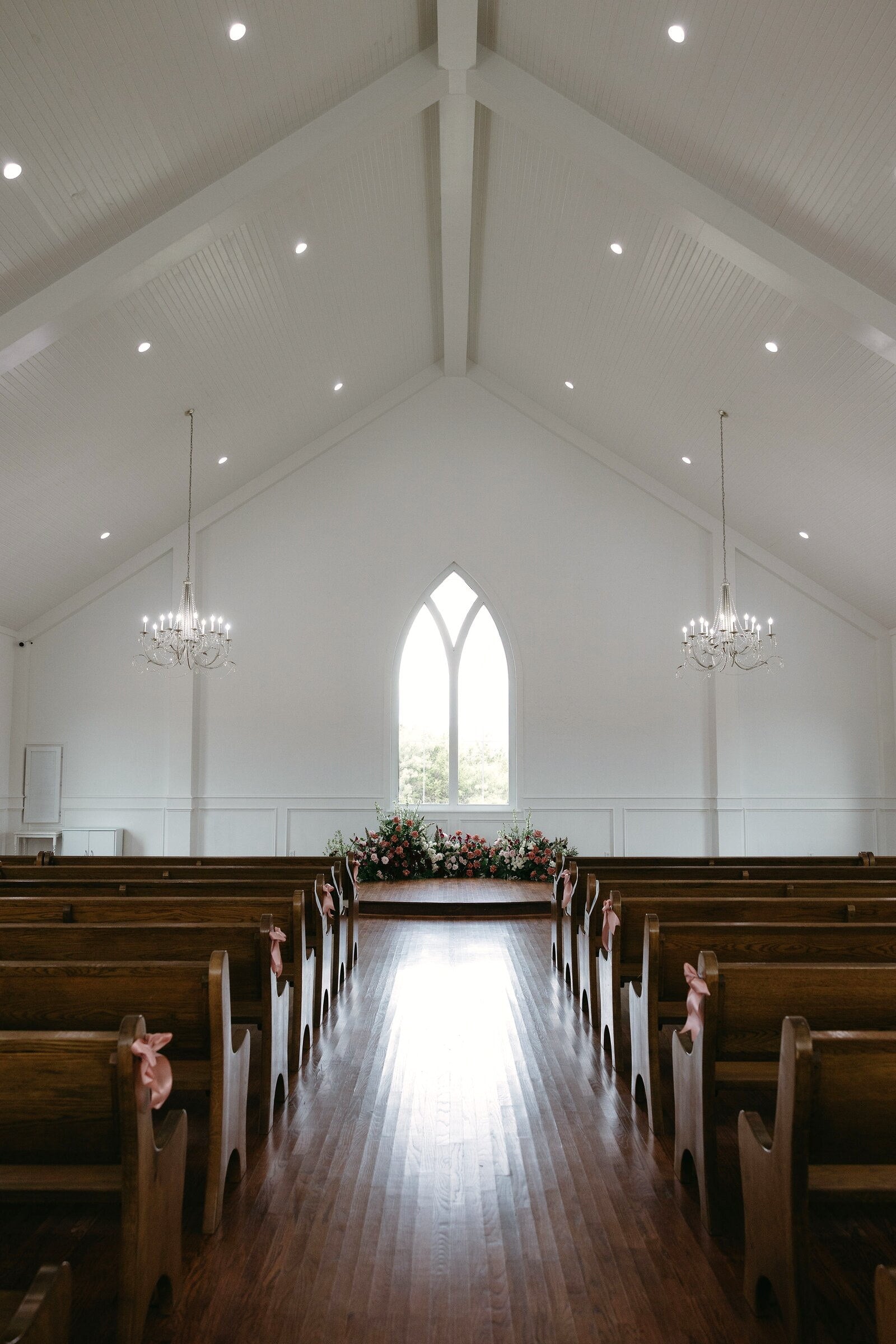 Symmetric view down the wood-pew aisle of The White Sparrow chapel to the arched window, blush aisle florals, by B.blumen