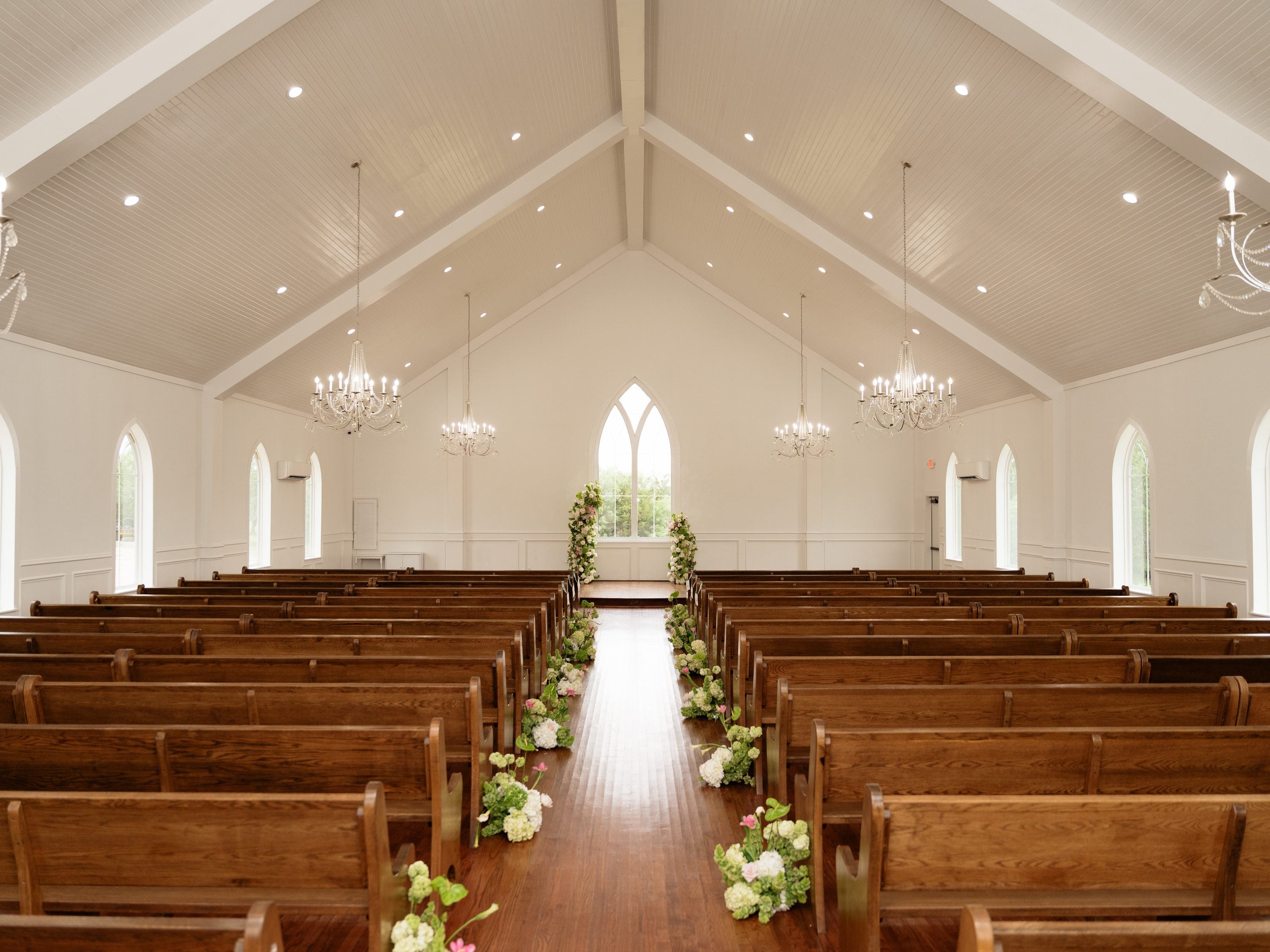 The chapel at The White Sparrow, wood pews under a vaulted white ceiling and chandeliers, white and green aisle florals running to the arched window at the altar, by B.blumen