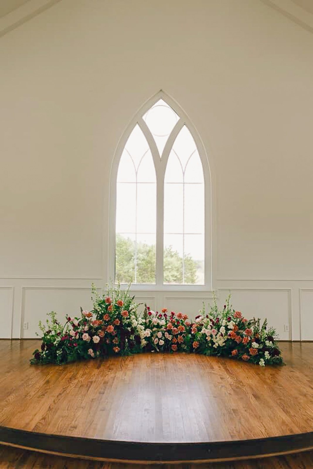 The arched chapel window at The White Sparrow with a low arc of pink and burgundy florals across the stage, by B.blumen