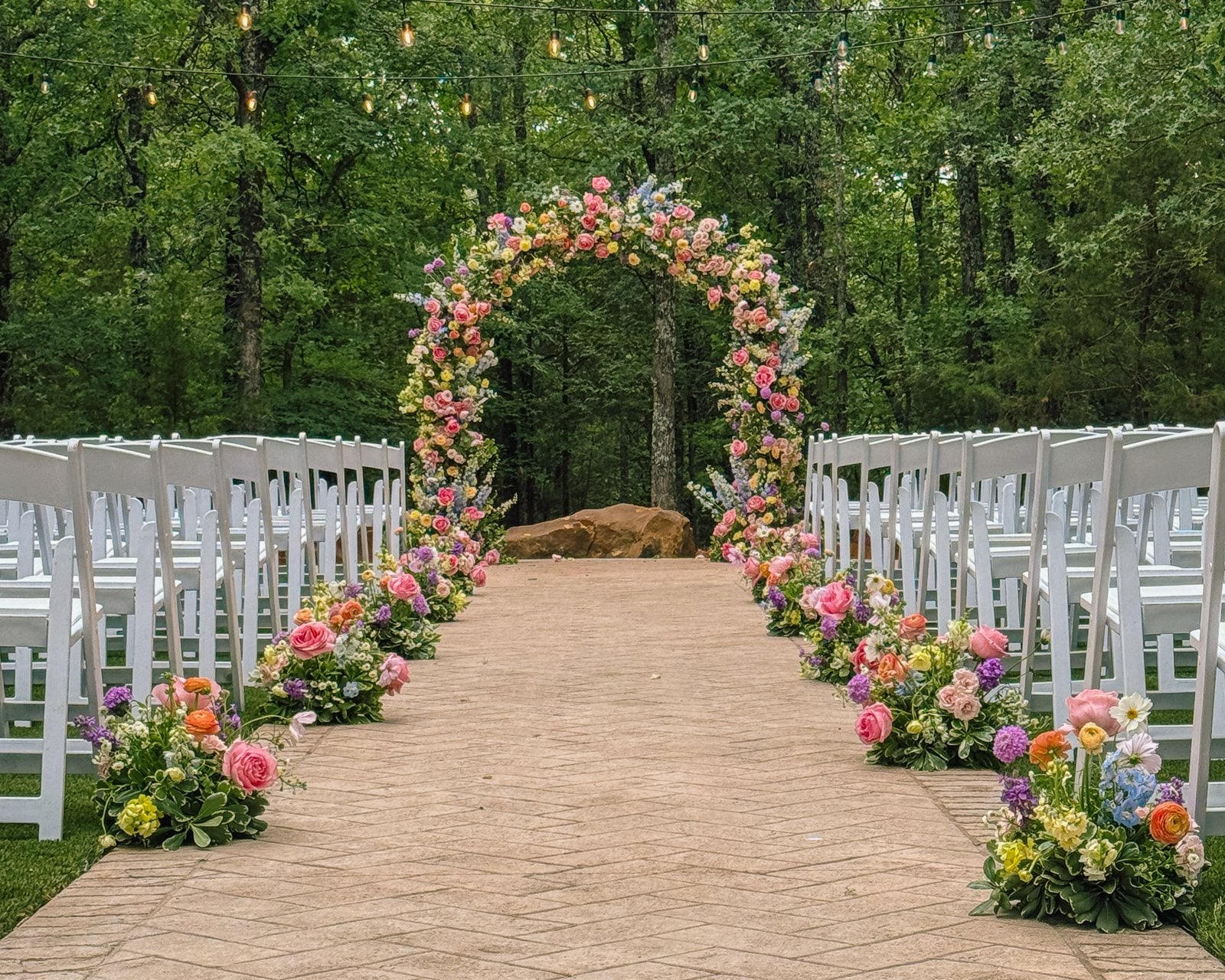 Wide view down the paved ceremony aisle at The White Sparrow to a full pastel floral arch, white chairs and aisle florals on both sides, trees behind, by B.blumen