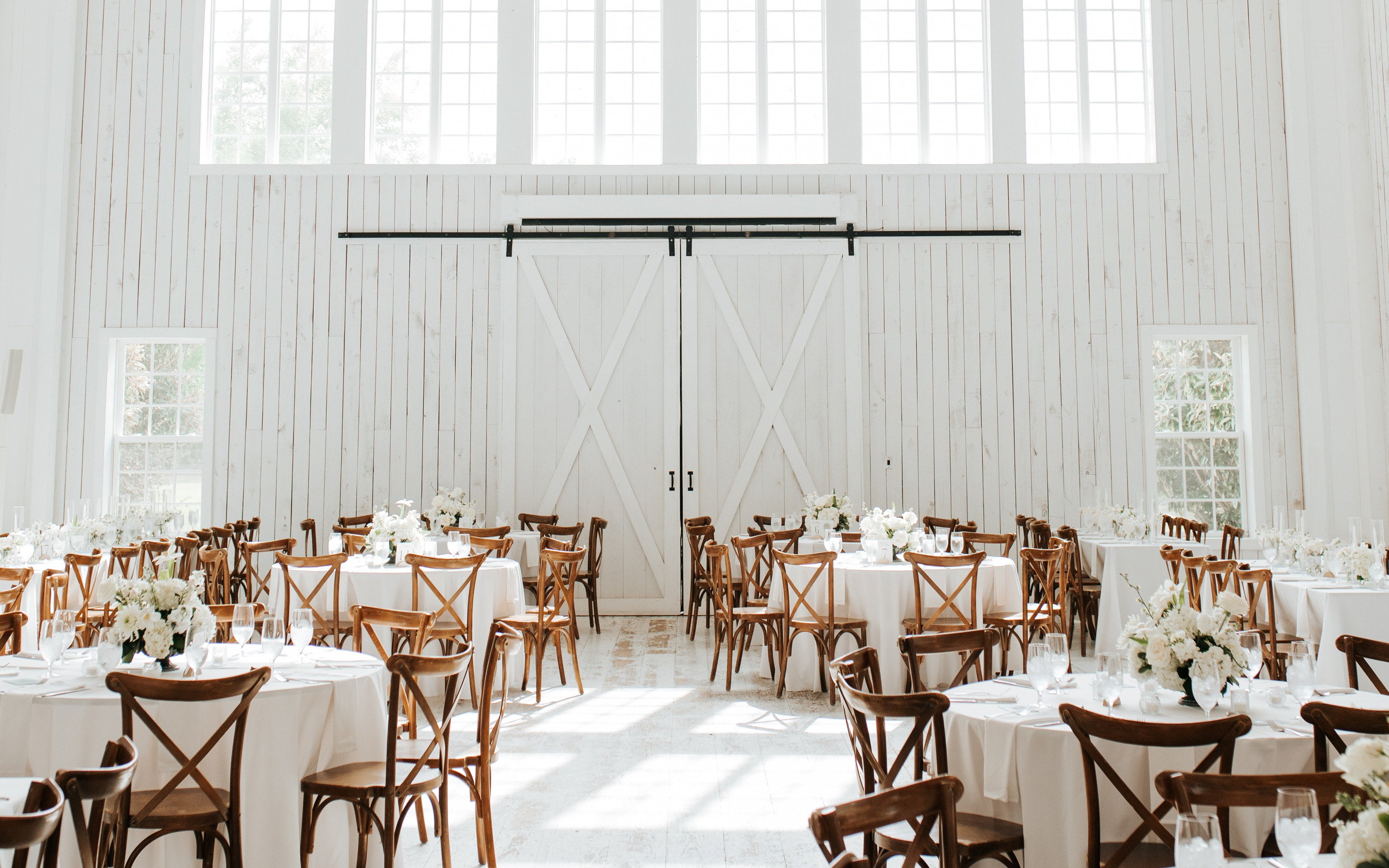 The White Sparrow hall set for dinner, round tables in white linen and cross-back chairs under the vaulted ceiling, the two-storey window wall and barn doors behind, by B.blumen