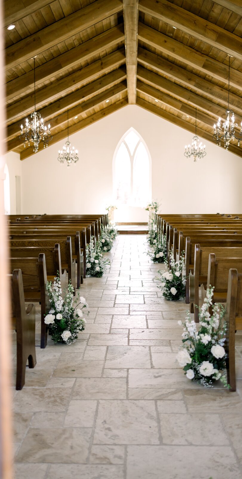 Interior of The Springs Waxahachie chapel with wooden pew aisle, gothic-arched window, and white floral aisle markers by B.blumen