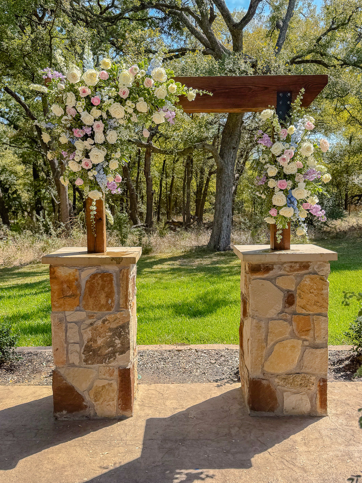 Two stone-pier pedestals at The Springs Alvarado topped with cascading pastel arrangements of white roses, blush, lavender, and pale blue blooms, beneath a wood-beam connector with the oak canopy behind, by B.blumen