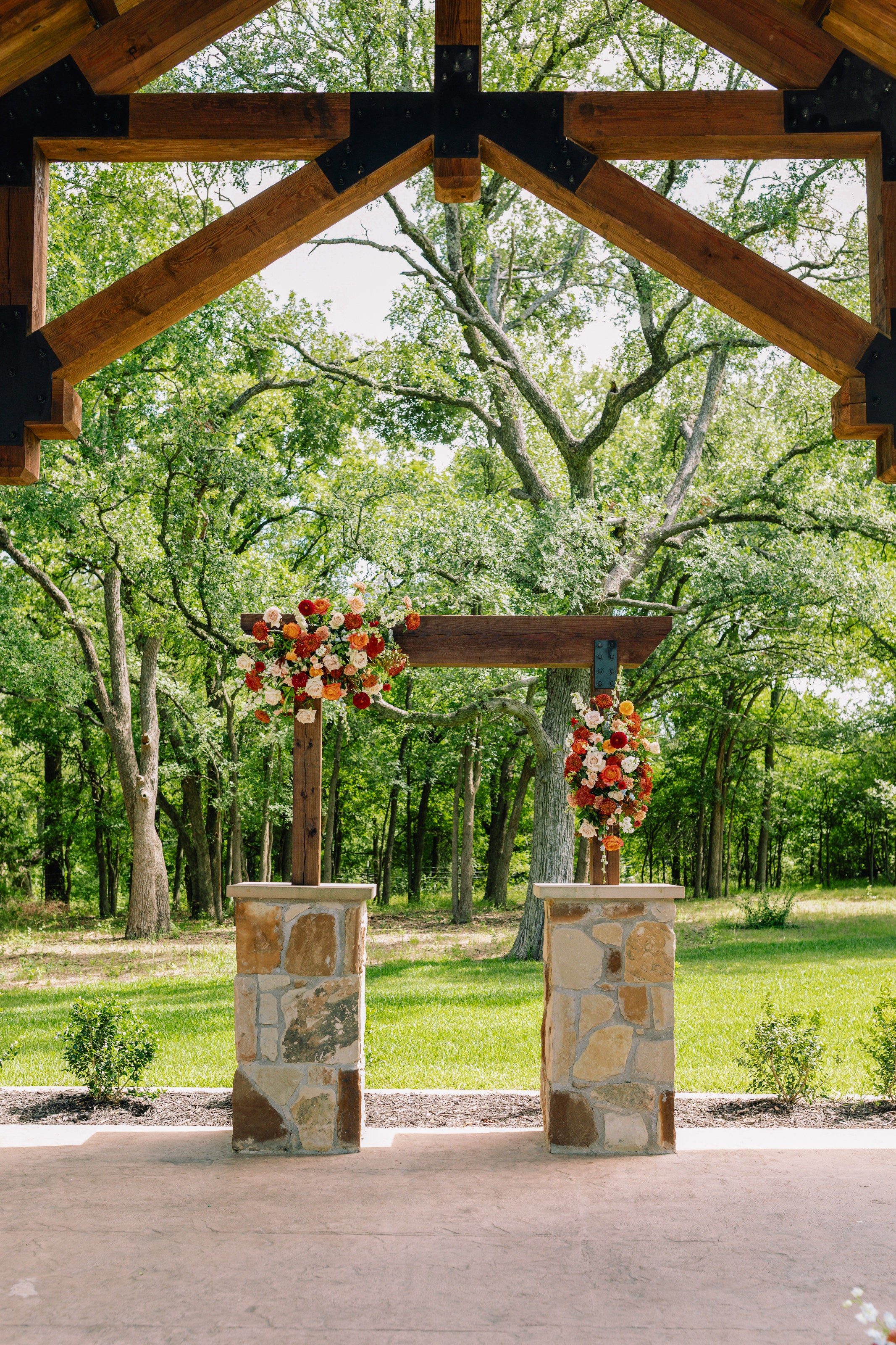 Ceremony arch under the wood-beam pergola at The Springs Alvarado, rust, orange and burgundy florals climbing the stone piers, by B.blumen